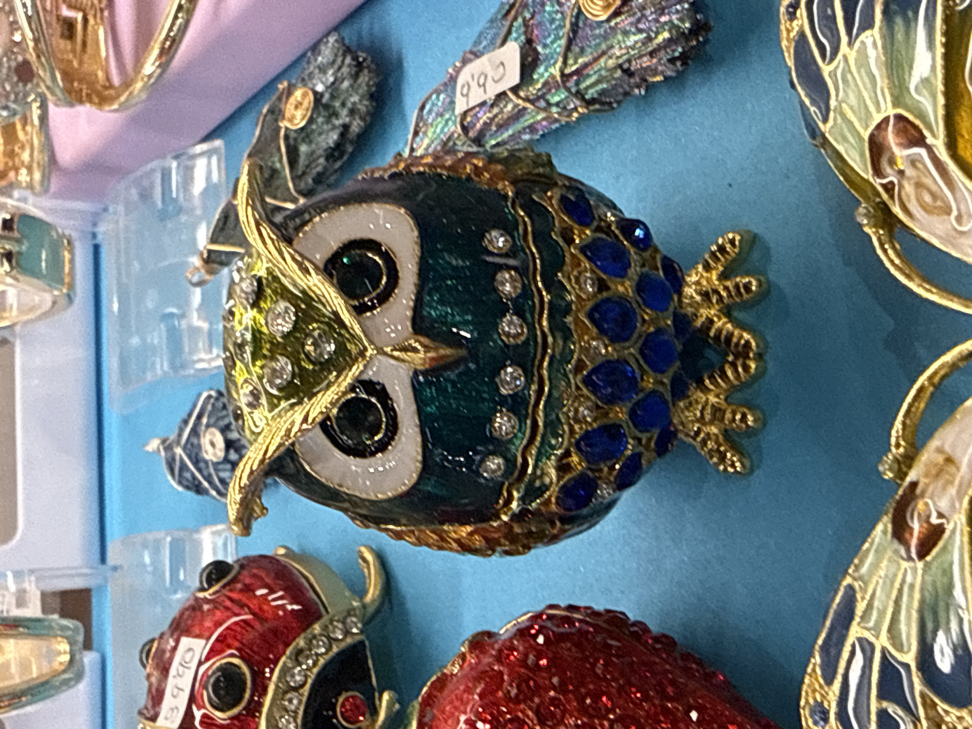 This close-up shot captures a vibrant display of ornate, jeweled trinket boxes, likely for sale in a gift shop or market in Puerto Del Carmen, Spain. The items are arranged against a light blue background, possibly a shelf or display surface.
The central and most prominent object is a large, elaborate owl-shaped trinket box. It lies horizontally, featuring a rich dark teal or green enamel body with intricate gold-colored metallic outlines and settings. Its surface is adorned with numerous sparkling clear rhinestones, particularly around its facial area and down its back, and larger royal blue rhinestones forming a pattern on one side. The owl has distinct white eyes with black pupils, also outlined in gold.
Above the owl, a smaller, dark, iridescent item resembling a bird or butterfly with shimmering abalone-like wings is visible, bearing a small white price tag that reads "9.95". To the right of the central owl, parts of two other butterfly-shaped trinket boxes with gold filigree and pastel enamel details are partially seen.
Below the main owl, a bright red, ladybug-shaped trinket box with black spots and clear rhinestone accents is visible, featuring a price tag that says "39.90". Further below and to the right, another red object with a glittery texture is partially shown.
The overall scene suggests a retail environment, with clear plastic risers visible on the left side of the frame, commonly used for product displays. The lighting appears even, suggesting an indoor setting during the day.