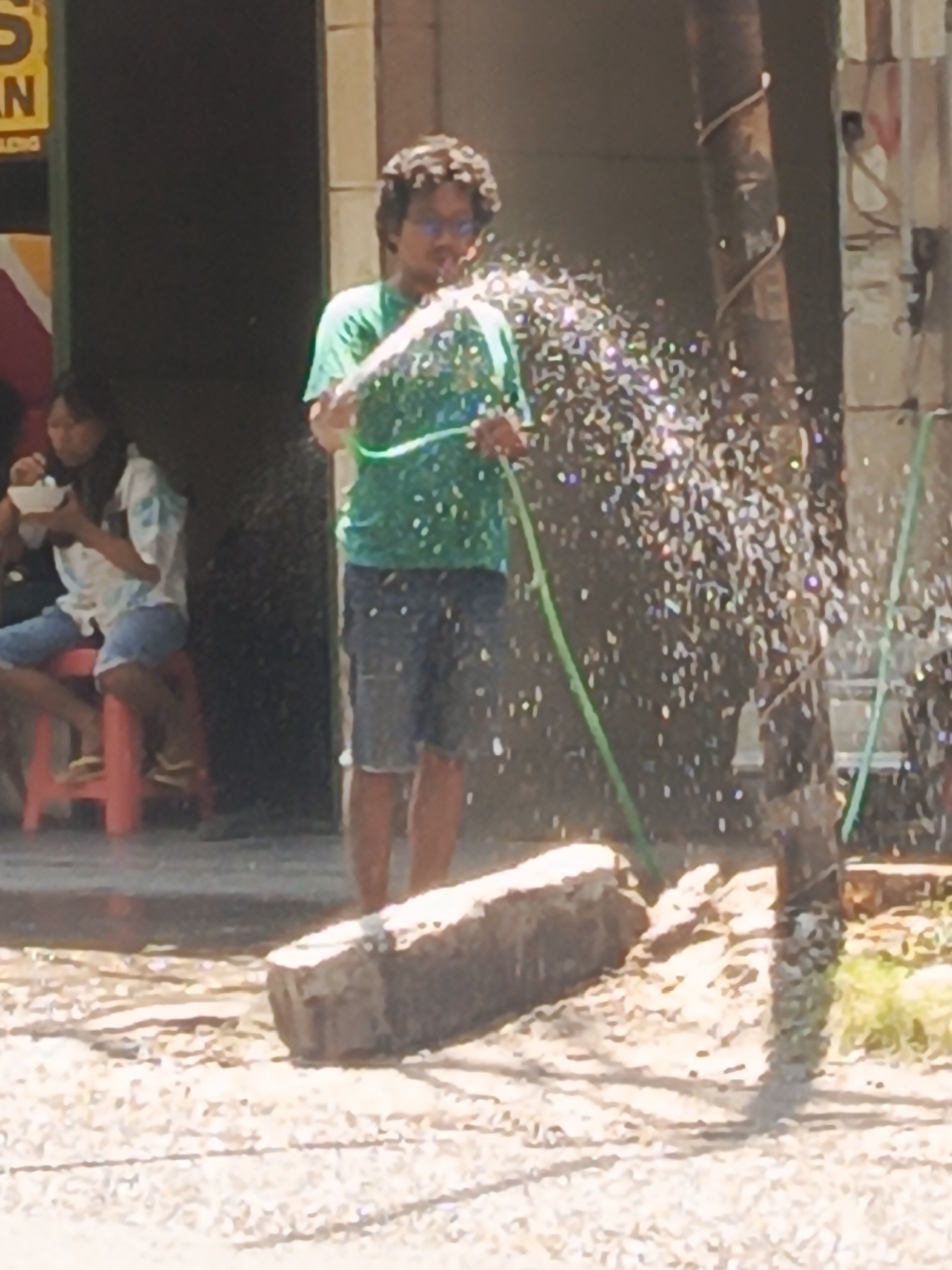 In Bungurasih, Indonesia, a young man with curly hair and glasses is spraying water from a green hose, creating a spray of droplets that are catching the light. He is wearing a green t-shirt and blue shorts. To his left, a young woman with dark hair is seated on a red stool, eating from a white bowl. She is wearing a white and blue patterned shirt and blue shorts. The background shows a dark doorway and a textured wall. In the foreground, a large, rough-textured rectangular object, possibly a concrete block, lies on the ground, which is covered in small stones and dirt. A thin, dark pole wrapped with a spiral design stands to the right of the man, with a green hose also visible near it. Green foliage is present on the right side of the image.