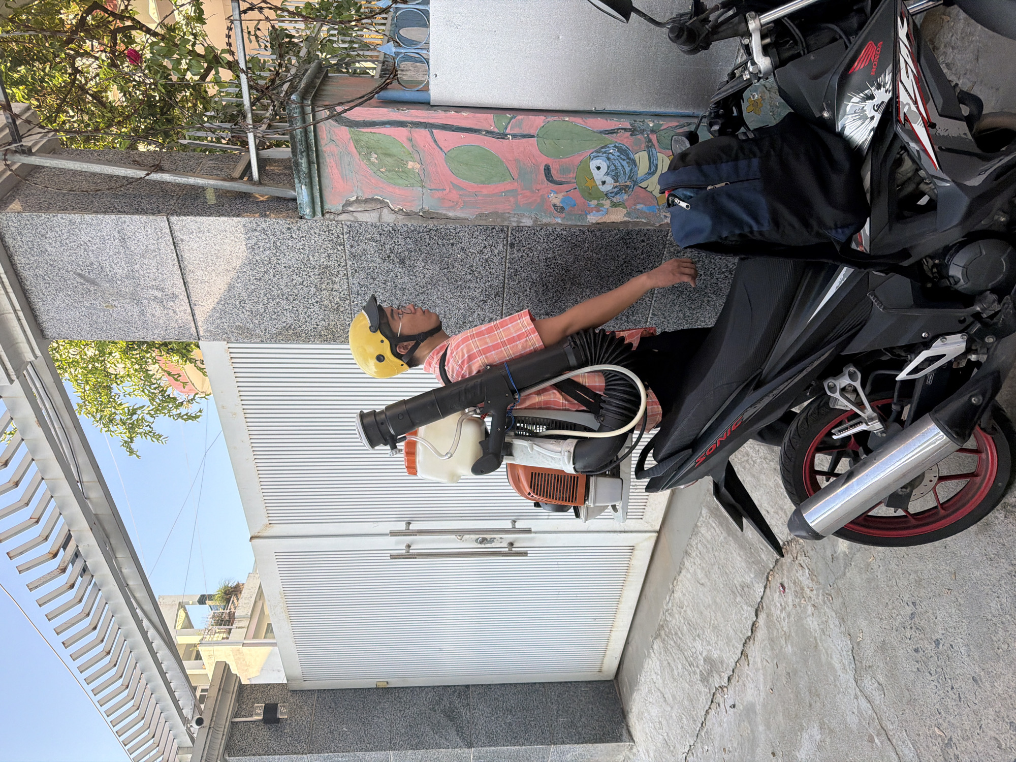 A person wearing a yellow helmet and glasses stands next to a motorcycle. They are also wearing a checkered shirt and dark pants. On their back, they are carrying a large spraying device. The motorcycle has a blue backpack on its seat and a red and black design on its side. The person is standing on a concrete path in front of a white metal gate and a granite pillar adorned with a colorful mural. Trees and a building are visible in the background. The scene appears to be a residential area in Bac Nha Trang Ward, Vietnam.