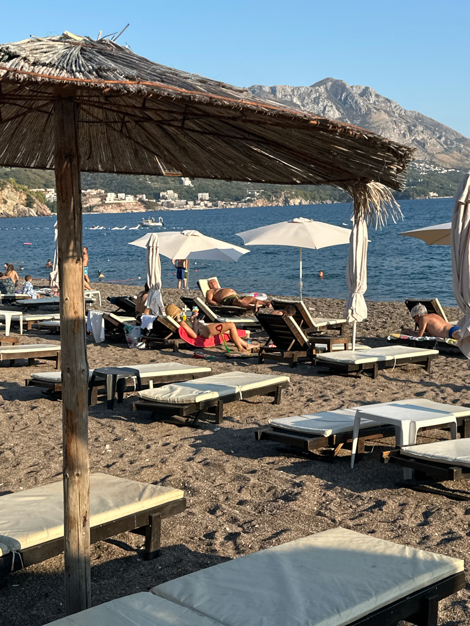 The image depicts a beach scene in Boreti, Montenegro, with the Adriatic Sea and mountains in the background. Several beach umbrellas, including a prominent straw one in the foreground, provide shade over rows of lounge chairs. People are scattered throughout the scene, some lounging on chairs, others walking or swimming in the water. A few boats are visible on the sea. The beach is composed of pebbles and sand. The sky is clear and blue.