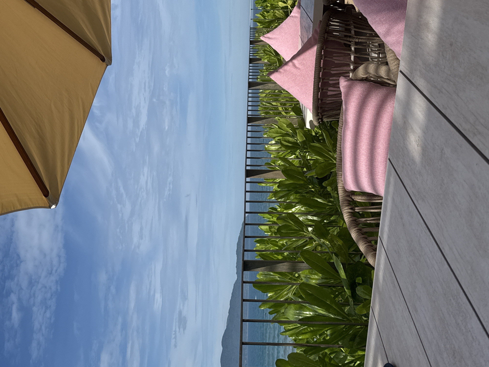 From a high-angle perspective, the image captures a serene coastal scene, likely in the city of Đà Nẵng, Vietnam. The foreground features a portion of a beige-colored parasol, extending into the upper-left corner of the frame. Adjacent to it, a patio area is furnished with rattan chairs adorned with rose-colored cushions. Lush green foliage provides a natural barrier between the seating area and a black metal railing. Beyond the railing, the scene opens up to a clear, blue sky speckled with wispy clouds, meeting the horizon of a calm, blue ocean. In the distance, a landmass is visible, suggesting a nearby island or peninsula. A patio table with a light grey surface is also visible in the foreground.