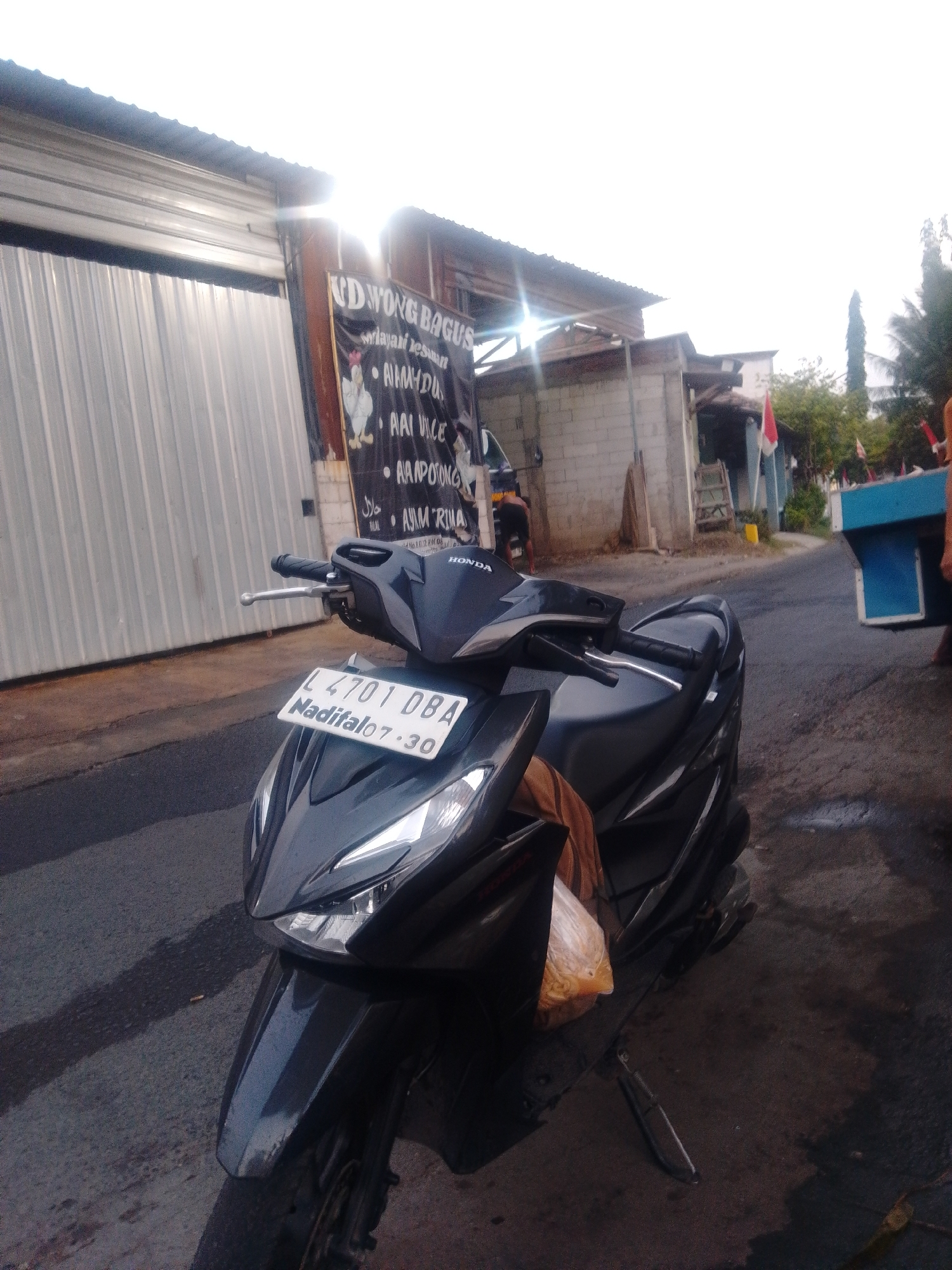 A dark, modern scooter is parked on a wet, asphalt road in front of a corrugated metal building with a partially opened roll-up door. A black banner with white text and images of chickens is hanging on the building's facade. The banner contains text such as "UD WONG BAGUS" and "MELAYANI PESANAN". The scooter's license plate reads "L 4701 DBA Maditalo 07.30". The sky is overcast with bright sunlight shining through from the left side of the frame. In the background, there are other buildings, including one made of concrete blocks, and some trees. An Indonesian flag is visible on one of the buildings. A person is partially visible in the distance to the right, near a blue cart. The scene appears to be a street in a commercial or residential area in Tambak Sumur, Indonesia.