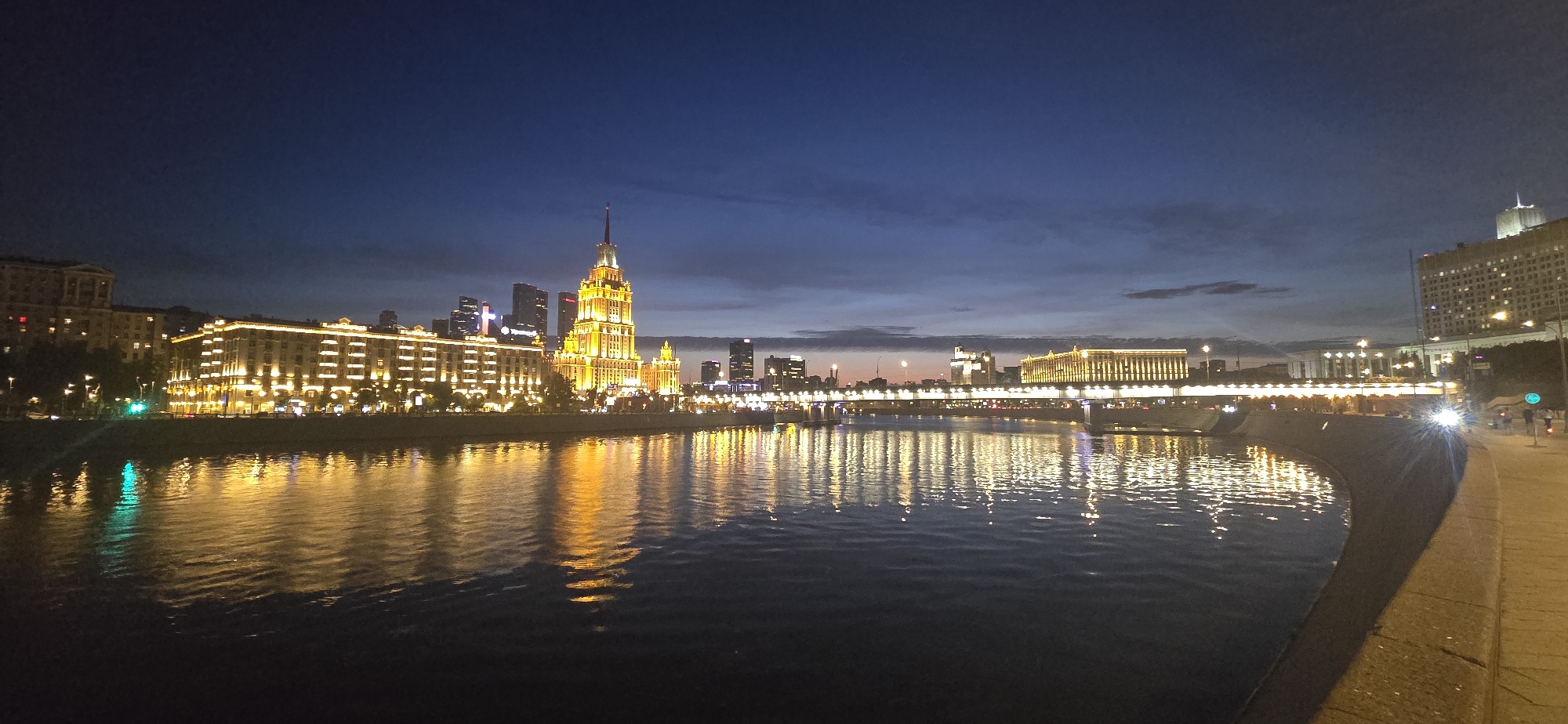 The image depicts a nighttime view of the Moskva River in Moscow, Russia. The riverfront is lined with illuminated buildings, including a prominent Stalinist skyscraper adorned with golden lights, which is characteristic of the Seven Sisters architectural style. The water reflects the city lights, creating a shimmering effect. Across the river, more buildings and bridges are visible, all lit up against the twilight sky. The scene suggests a vibrant urban environment at night.