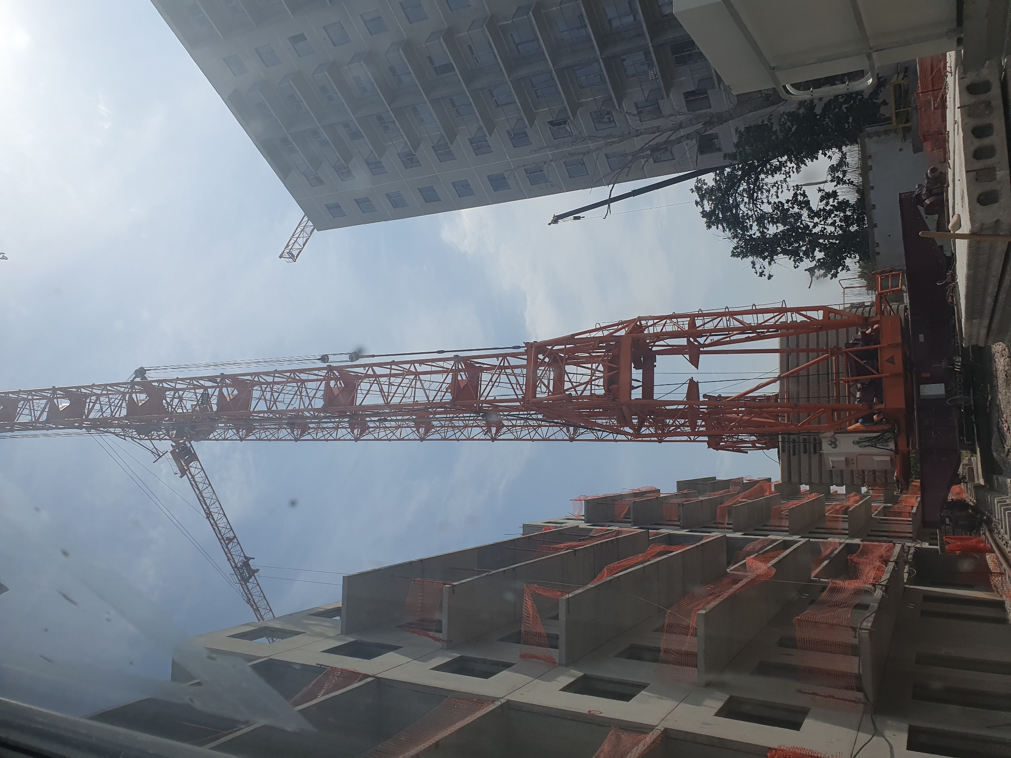 An exterior, low-angle, slightly upward-facing, full shot captures a construction scene on a bright, partly cloudy day. Dominating the frame is a tall, orange construction crane with a lattice structure. To the left, a multi-story concrete building under construction is visible, featuring open floors and orange safety netting on balconies. To the right, another, taller, grey apartment building with multiple floors and balconies is partially obscured by the crane. In the foreground, concrete slabs are stacked on the ground. A smaller, dark boom crane is visible to the right of the large orange crane, positioned behind the grey apartment building. The scene appears to be in a developing urban area, likely Tashkent, Uzbekistan, given the architectural style of the buildings. No individuals are clearly discernible in the image.