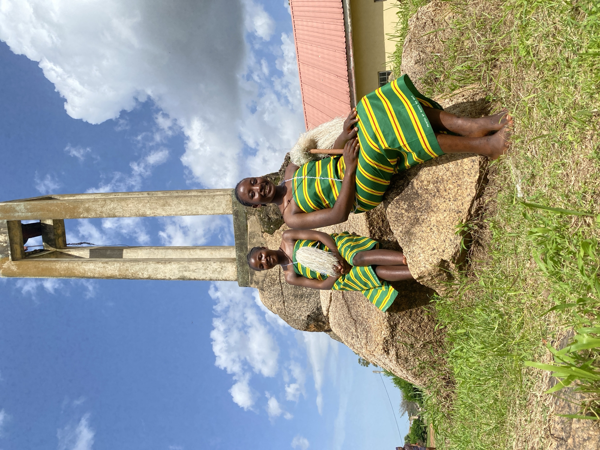 In Dutsen Kato, Nigeria, two young girls are captured in a posed photograph against a backdrop of a bright blue sky with scattered white clouds. The image is oriented vertically, with the subjects positioned on a large, weathered rock formation. Both girls are dressed in vibrant, traditional attire featuring green and yellow stripes with thin red accents. The fabric has visible text indicating it was "PRINTED BY SUPER MK.K."
The girl in the foreground, seated higher on the rock, is looking towards the camera with a warm, engaging smile. She wears a necklace and holds a fluffy, white object in her hands, possibly part of a costume or cultural accessory. Her legs are crossed, and her bare feet rest on the rock. The second girl is positioned slightly behind and below her, her face partially obscured but also appearing to be engaged with the camera. She also wears the same striped outfit and holds a similar white accessory.
To the left of the frame, a large concrete structure with visible rebar rises towards the sky, suggesting the presence of a building or tower nearby. A portion of a red-roofed building is visible in the upper right of the image, beyond the rock formation. The ground around the rock is covered in dry grass, indicating a potentially warm or dry climate. The lighting suggests it is daytime, with the sun casting subtle shadows. The overall mood of the image appears cheerful and celebratory, possibly capturing a cultural event or a moment of joy.
