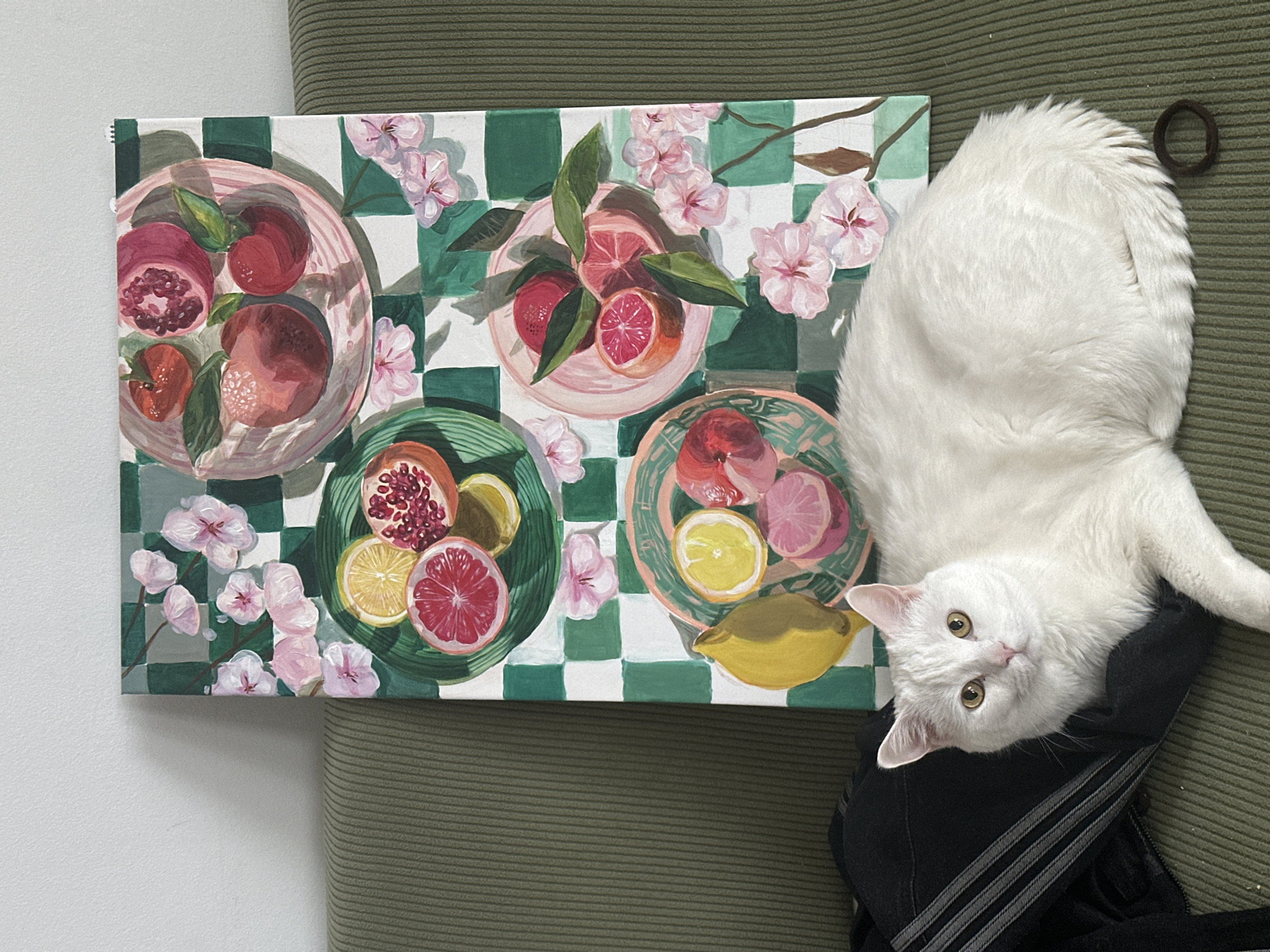 The image shows a painting of fruit on plates arranged on a checkered green and white tablecloth with flowering branches lying next to a fluffy white cat with yellow eyes, sitting atop a black Adidas tracksuit on an olive green corduroy surface. The location is not discernible based on the image provided.