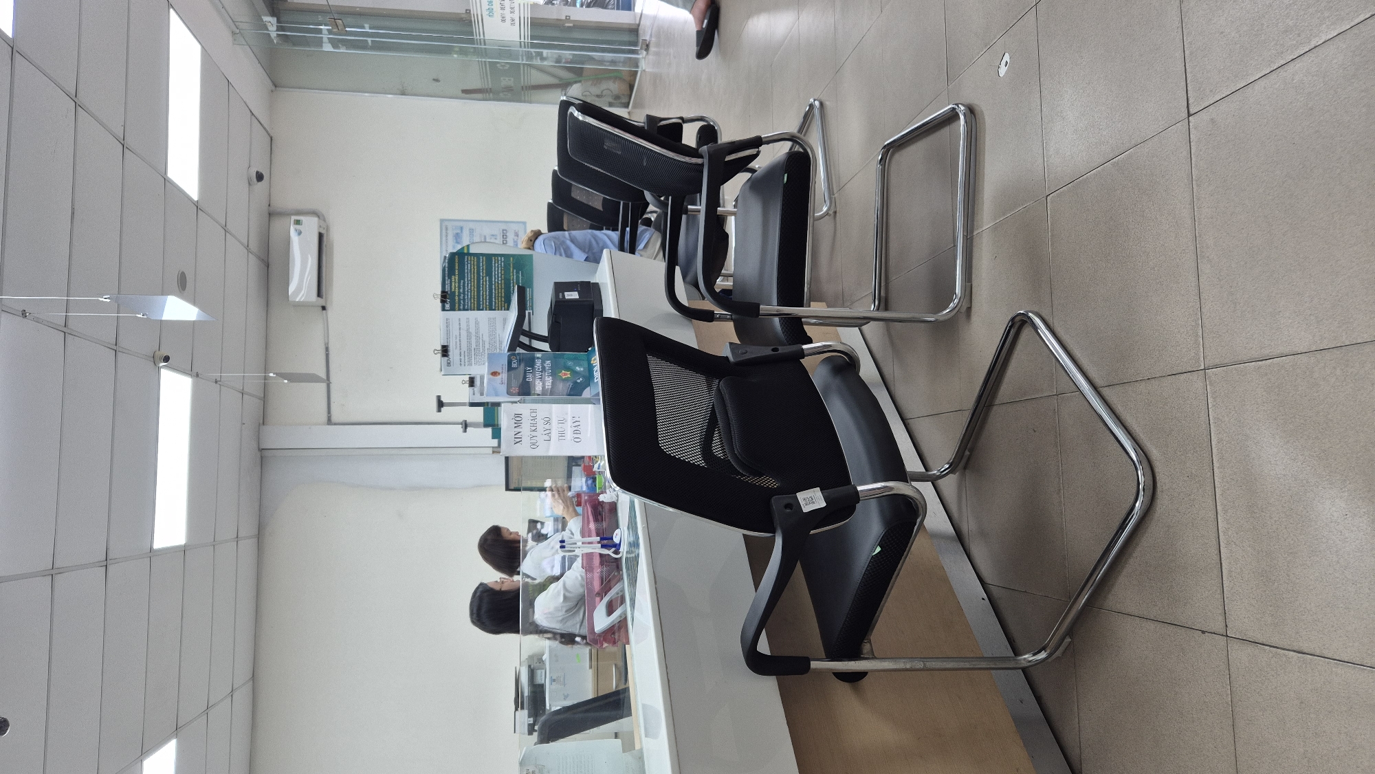 This image depicts the interior of a bank branch in the city of Da Nang, Vietnam. In the foreground, two black mesh office chairs with chrome sled bases are positioned on a tiled floor. The chair on the left is closer to the viewer and has a QR code sticker on its left leg. Behind these chairs, a white counter with a glass partition separates the customer area from the staff area. On the counter, there are various items including a printer, a scanner, and a sign with Vietnamese text that reads "XIN MỜI QUÝ KHÁCH LẤY SỐ THỨ TỰ Ở ĐÂY!" which translates to "CUSTOMERS ARE INVITED TO GET YOUR NUMBER HERE!".
Two female employees are visible behind the counter, working on computers. The employee on the left is wearing a white shirt and looking at her computer screen. The employee on the right, also in a white shirt, is holding a pen and appears to be interacting with documents or a customer. A third person, wearing a blue shirt, is partially visible in the background, seemingly assisting another customer. The environment is brightly lit with overhead fluorescent lights, and an air conditioning unit is mounted on the wall. The overall scene suggests a typical banking service environment.