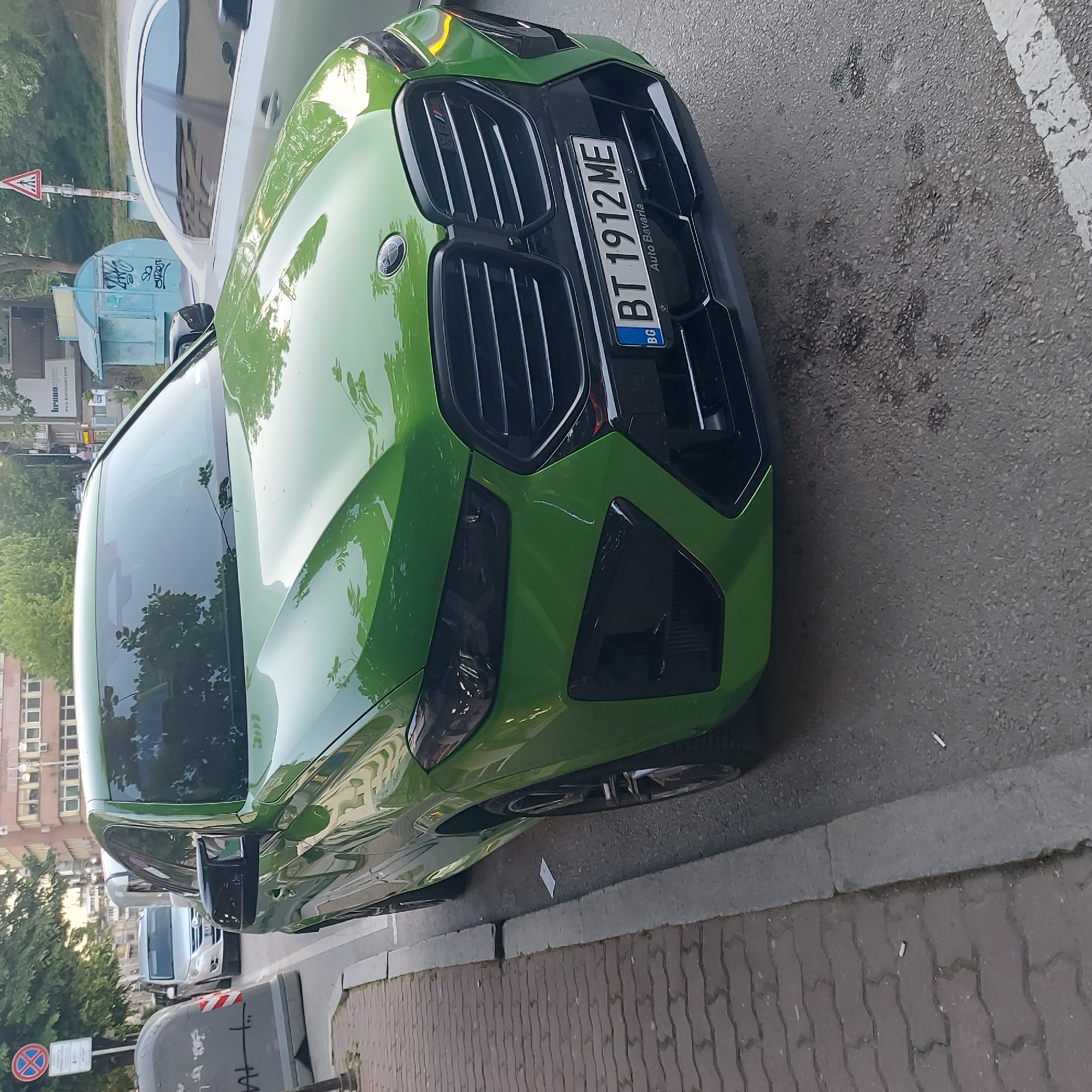 The image shows a green BMW parked on the side of a street. The license plate number of the car is BT 1912 ME. The car is parked on a street in what appears to be a European city. Based on the license plate, the car is from Bulgaria. No people are visible. The specific location within the city of Veliko Tarnovo is not evident from the image. The background features buildings and trees. A white vehicle is parked in front of the BMW.