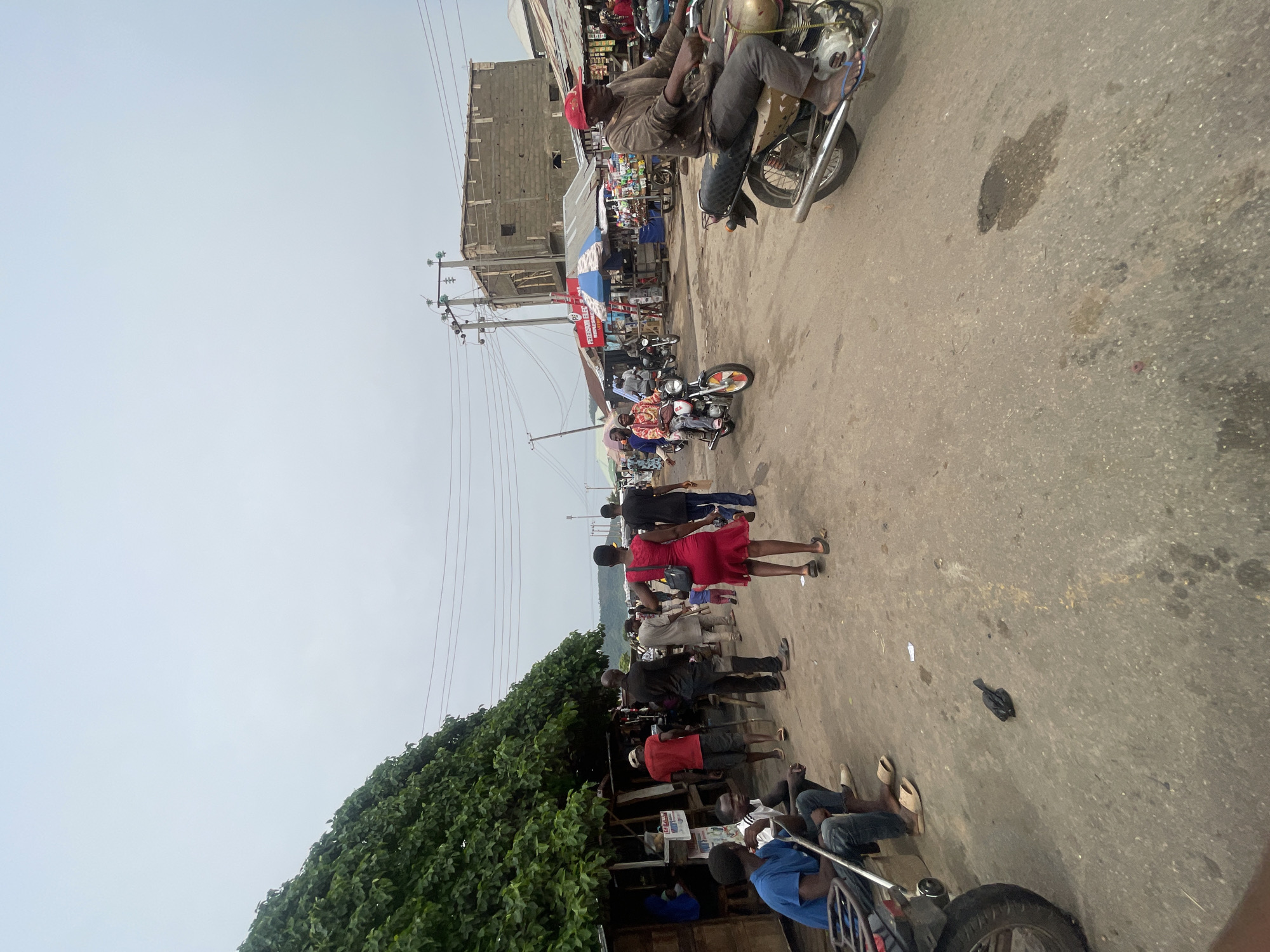 The image depicts a street scene in Lokoja, Nigeria. A group of people is walking along a paved road. There are parked motorcycles, some occupied, along the side of the road and a person is riding a motorcycle at the top of the frame. Buildings and vendors with awnings are visible in the background. A large tree is on the left side of the frame. The sky is overcast. There are electrical lines hanging overhead.