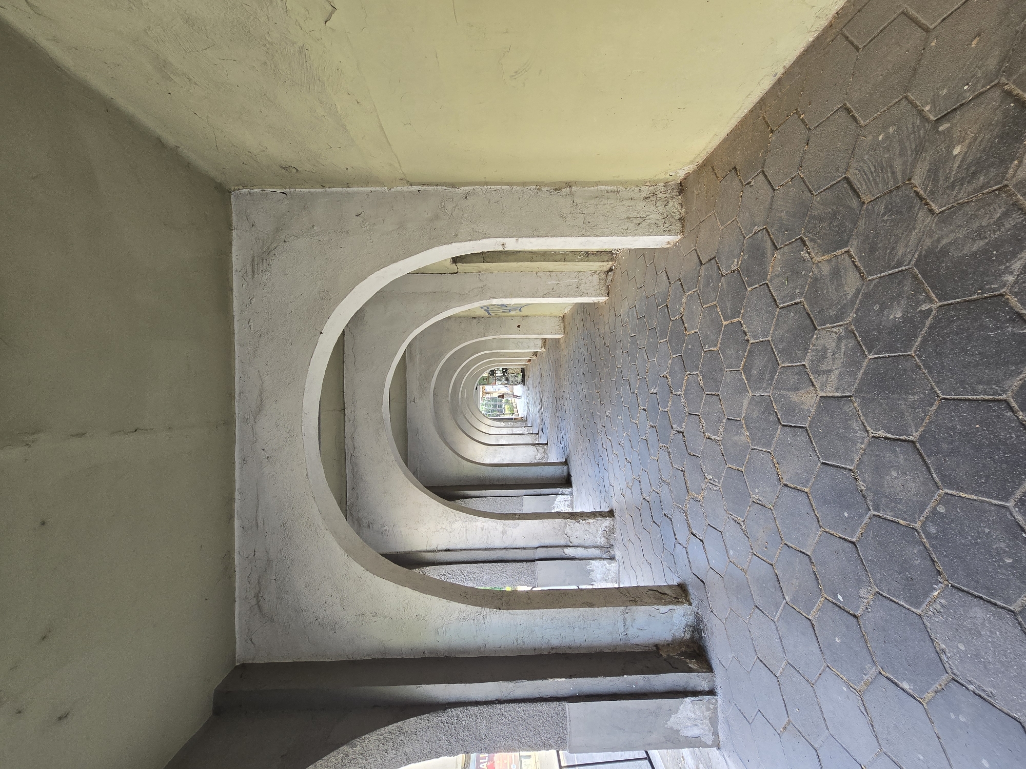 A perspective shot looking down a corridor of repeating concrete arches. The arches are spaced evenly and are connected at the top to a yellow-painted ceiling. To the right of the arches is a floor of hexagonal pavers in a dark gray color. The corridor leads to an opening at the far end, revealing a glimpse of what appears to be a street with trees and buildings. The light increases gradually down the corridor. The location appears to be outdoors, possibly an underpass or a covered walkway. There are no people visible in the shot.