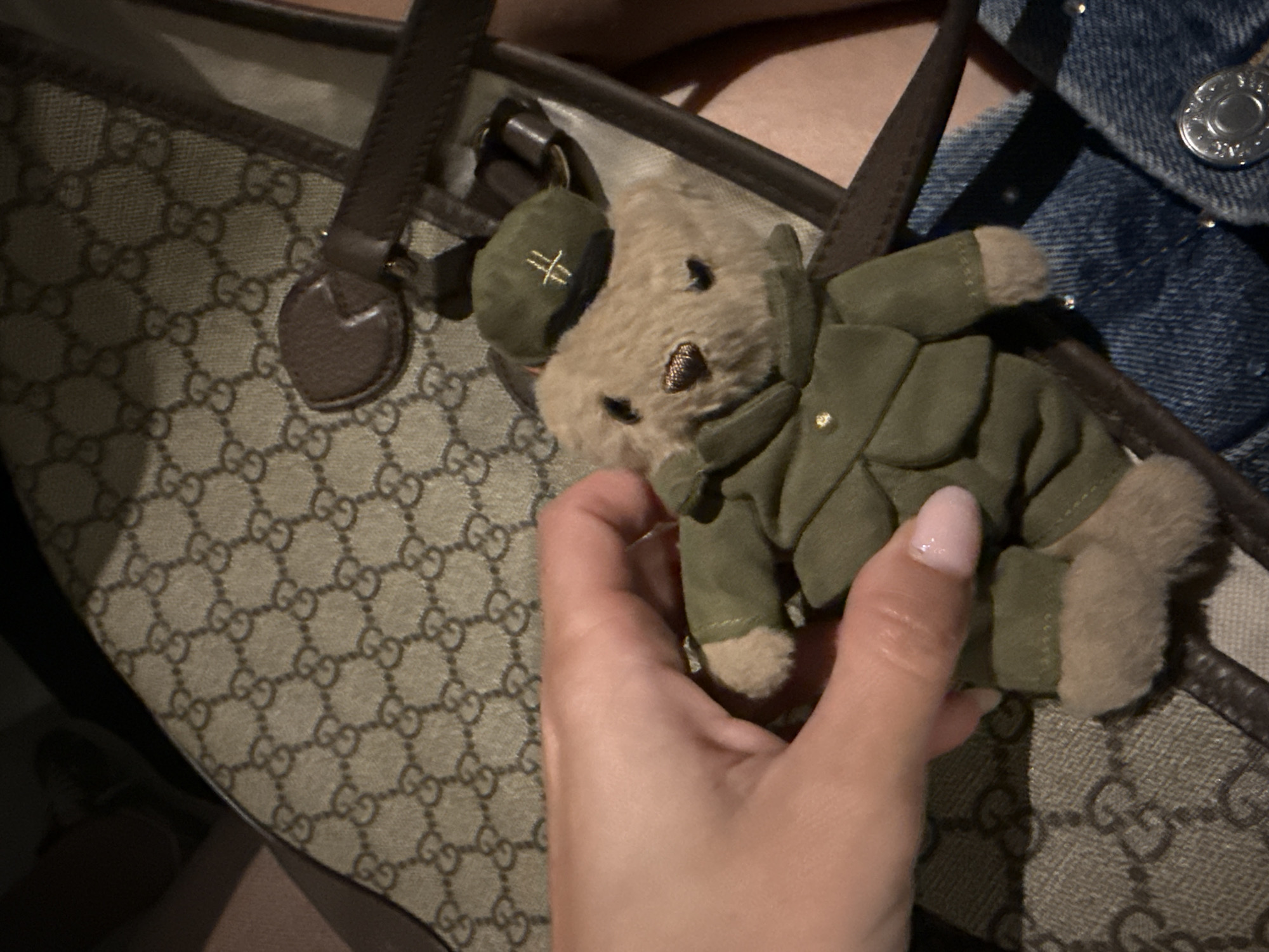The image displays a close-up, downward-angled view of a small teddy bear held by a human hand, set against a luxury handbag and denim fabric. This scene is located in Cape Town, South Africa. The teddy bear is light brown and dressed in an olive-green military-style uniform, consisting of a jacket with small gold-colored buttons and a matching beret featuring a gold embroidered symbol. A human hand, with light skin and manicured nails bearing pink polish, gently holds the teddy bear by its torso. The primary background object is a light brown Gucci monogram canvas bag with dark brown leather trim and strap detailing. In the lower right quadrant, blue denim fabric with a silver-colored metal button is visible, indicating the clothing worn by the individual holding the bear.