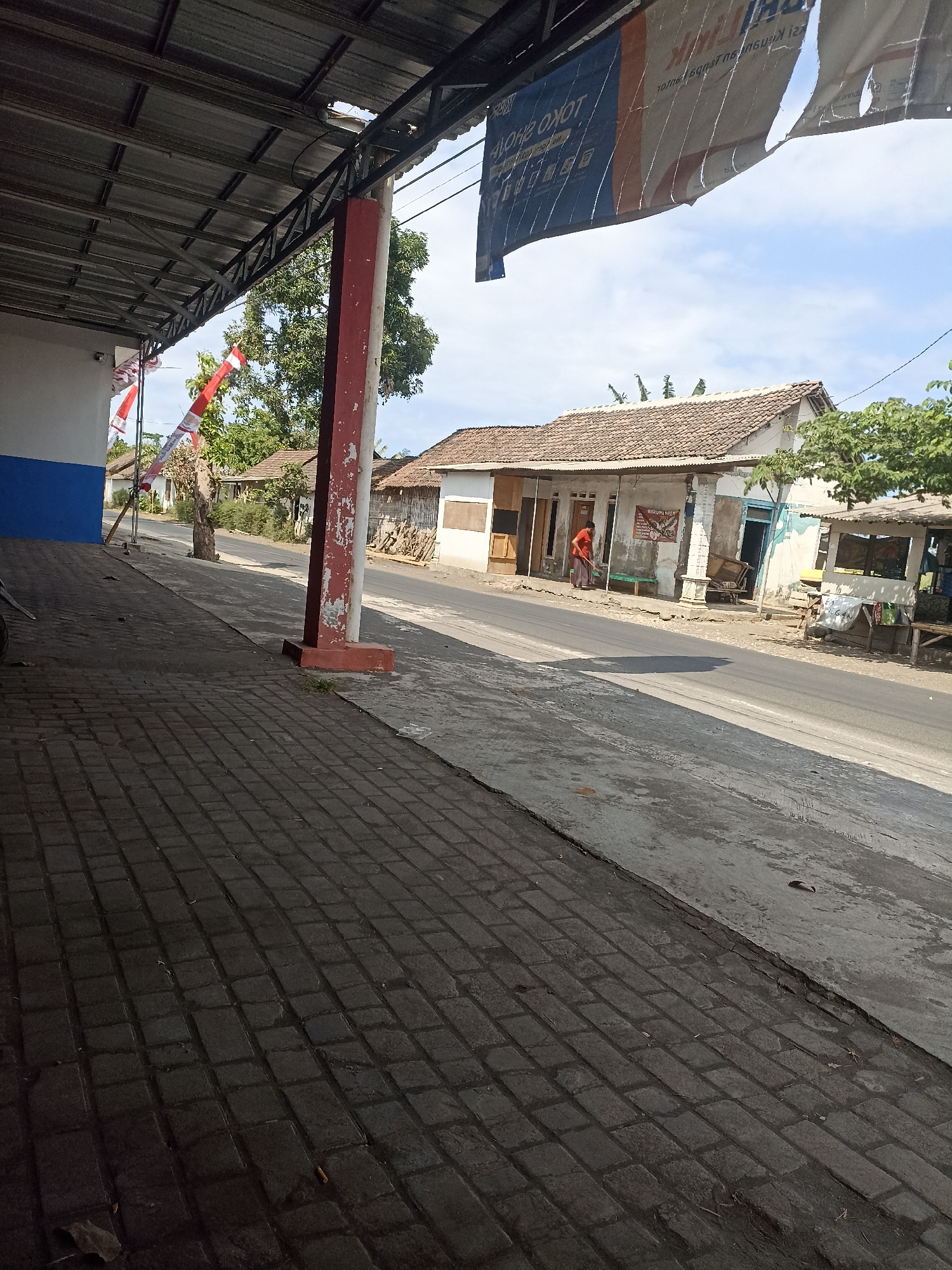This is a vertical, eye-level, medium-wide shot of a street scene in Pandanarum, Indonesia. The foreground is dominated by a textured, dark grey brick sidewalk that slopes gently upwards towards the right. To the left, a portion of a building with a blue and white facade is visible, with a metal overhang extending over the sidewalk. A red-and-white Indonesian flag is attached to a pole near the building.
The middle ground features a paved road running parallel to the sidewalk. On the opposite side of the road, a row of modest, single-story buildings with tiled roofs line the street. One building appears to be a shop, with a person in an orange shirt sweeping the area in front of it. A wheelbarrow and other items are visible near this building. Further down the street, more buildings and trees are present under a bright, partially cloudy sky. A tattered banner hangs from the metal overhang in the upper portion of the image, partially obscuring the sky. The overall impression is that of a quiet, rural or semi-urban street on a sunny day.