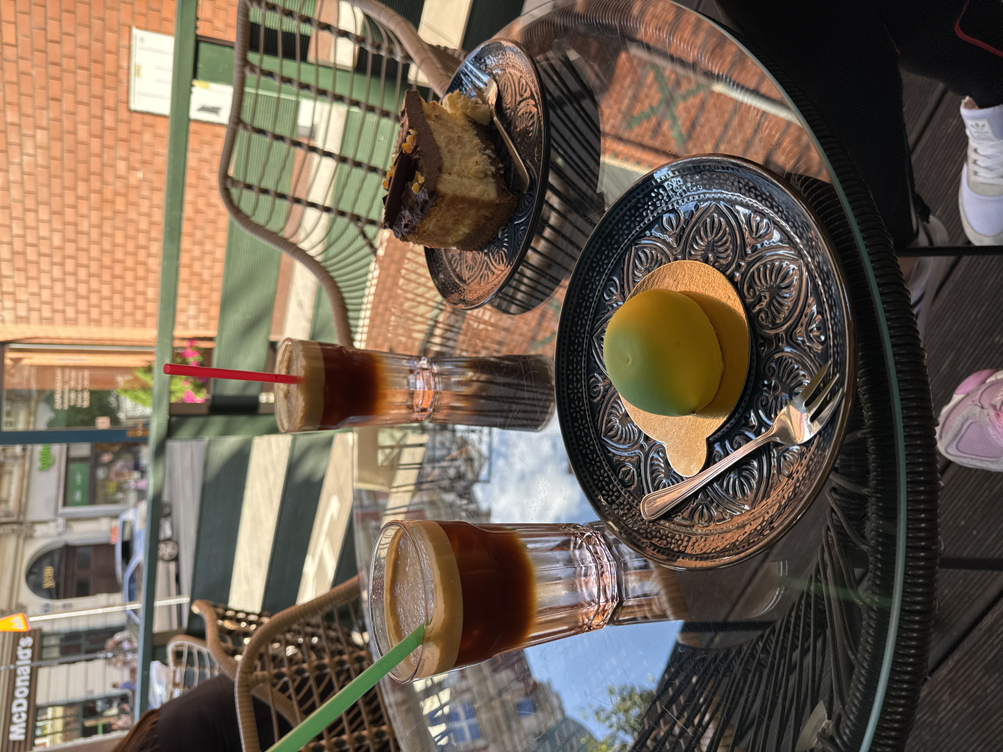 This image captures an outdoor cafe scene on a sunny day in an unknown city. A round glass-top table is centered, reflecting elements of the background and surroundings. On the table, two tall, clear glasses hold layered cold beverages, likely coffee, with a dark brown liquid bottom and a lighter, foamy top. The glass on the left has a green plastic straw, while the one on the right has a red plastic straw and is partially obscured. Two desserts are also on the table: on the left, a small, spherical, light green pastry rests on a gold foil base atop a dark blue/black ceramic plate with an intricate embossed pattern; a small silver fork is beside it. On the right, a slice of cheesecake with a dark chocolate topping and orange zest decoration is presented on an identical dark ceramic plate, with another small silver fork inserted into the cake.
Partially visible wicker-style chairs surround the table. To the far left, a person's arm wearing a dark top is visible. Beneath the table, two pairs of feet are visible on a dark wooden slatted floor: one foot wears a light pink and white sneaker, and the other wears a white sneaker.
In the background, beyond a dark green horizontal railing, a street scene unfolds. Buildings with light-colored facades and windows line the street, including a prominent "McDonald's" sign on one building. A light-colored car is visible on the street, and several blurry figures of pedestrians can be seen walking. Some green foliage and pink flowers are visible behind the railing. On the far right background, a reddish-brown brick wall with white utility boxes is present. The scene is illuminated by bright daylight, casting reflections on the glass table.