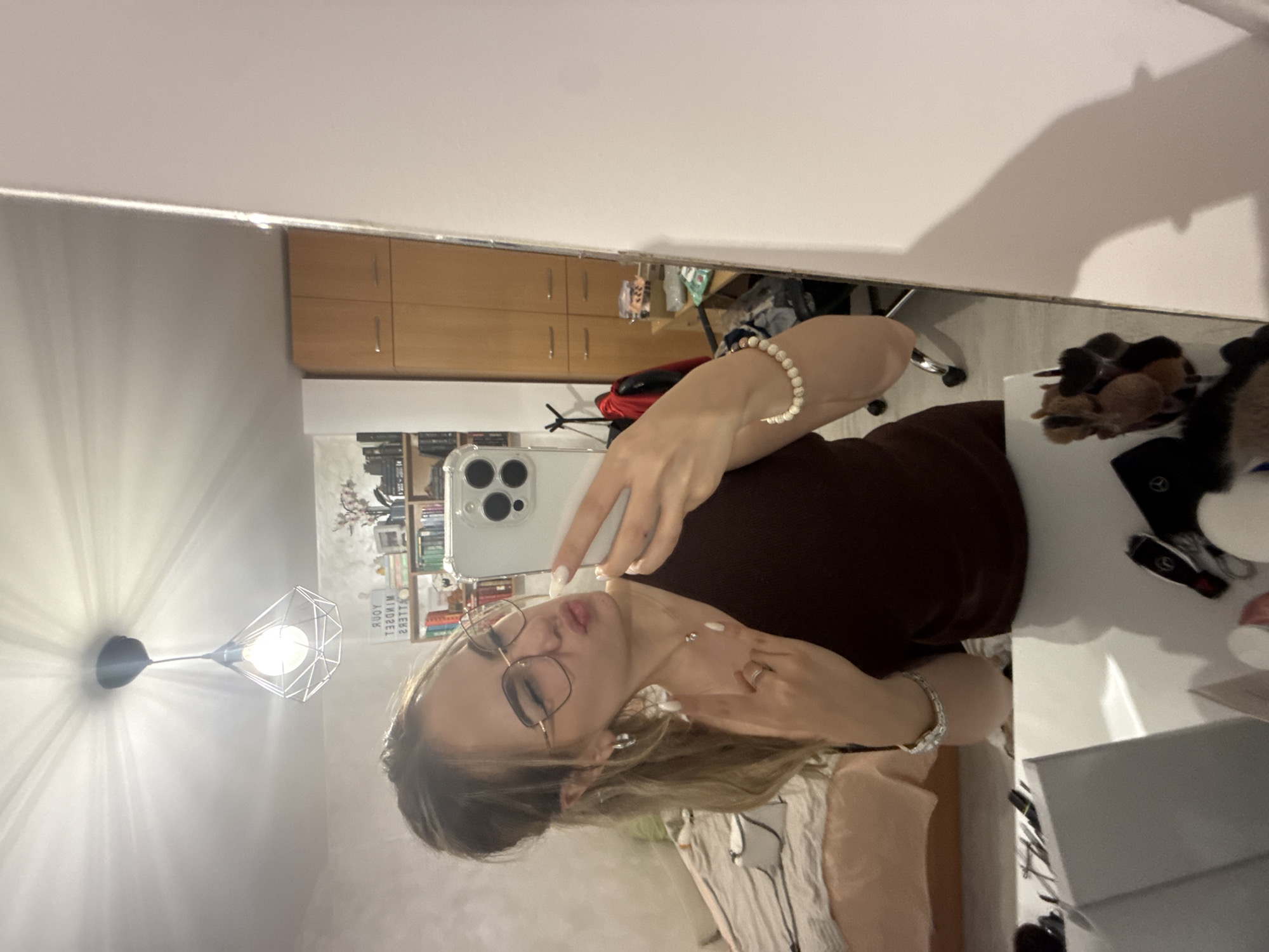 The image is a selfie taken in what appears to be a private indoor setting, possibly a bedroom or apartment. The photographer is a young woman with blonde hair, wearing glasses, a brown top, and jewelry (necklace and bracelet). She is holding a phone (iPhone) in front of her, presumably to capture the image in a mirror. The reflection in the mirror reveals parts of a room with bookshelves, a lamp fixture on the wall, and wooden cabinets. There is a desk or table with various objects, including what seems to be a makeup brush set, car keys, and other personal items. The room's decor suggests a modern or contemporary style. There is no indication of the location being in the city of Blagoevgrad, Bulgaria, or any other specific geographic area.