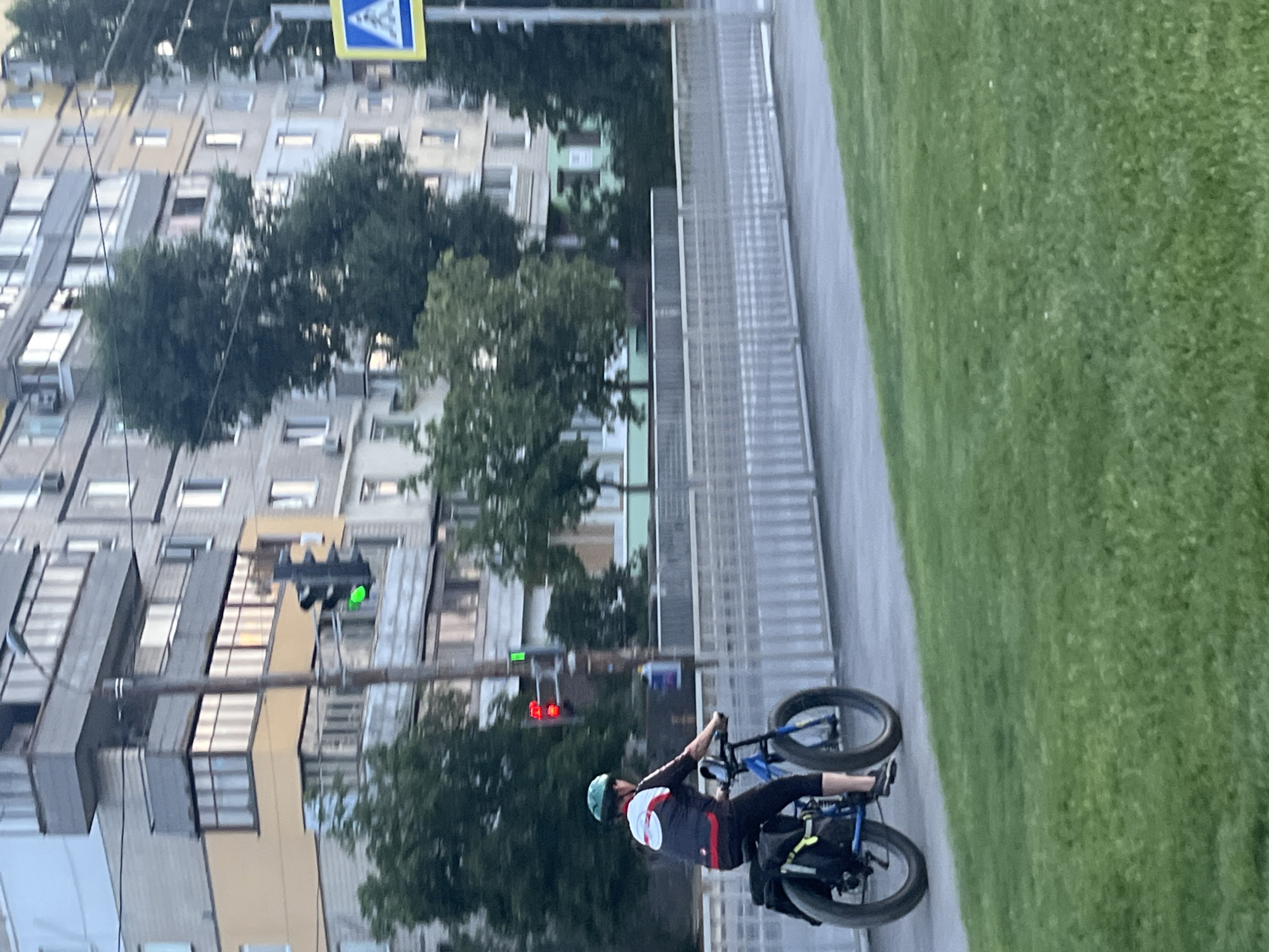 An outdoor, eye-level shot captures a person riding a blue fat-tire bicycle on a paved path. The individual, wearing a green helmet, a dark cycling jersey with white and red accents, and black leggings, is positioned in the lower-left quadrant of the frame, facing right. The bicycle has black saddlebags attached to the rear rack. Behind the cyclist, a grey metal fence runs horizontally across the mid-ground. In the background, a multi-story apartment building with balconies and windows is visible, with trees interspersed. A traffic light is mounted on a pole in the mid-ground; its top light is red, and its bottom light is green. A pedestrian crossing sign with a yellow background and a black pedestrian symbol is located on the far right, on a pole. The foreground is a grassy lawn. The scene is set in the city of Dnipro, Ukraine.