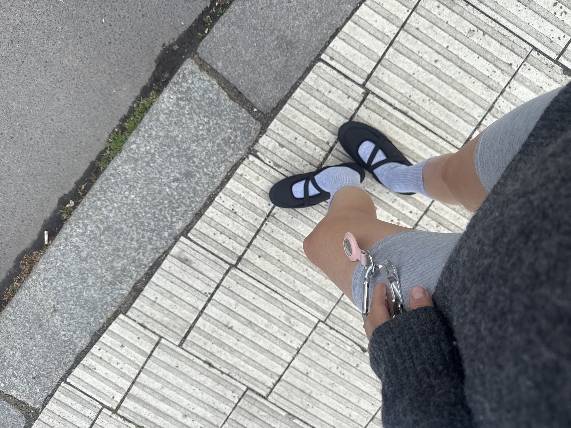 A person is standing on a sidewalk made of textured, off-white rectangular tiles, with black shoes and light gray socks. The person is wearing light gray shorts and a dark gray sweater, and holding a silver nail clipper with a pink attachment in their right hand. To the left of the tiled sidewalk is a curb and a section of dark gray asphalt. The photo was taken in Berlin, Germany.