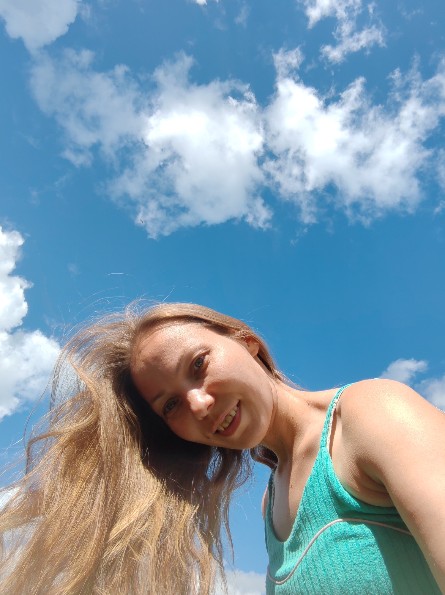 A low-angle, close-up shot shows a young woman looking up towards the camera, her face framed by long, blonde hair blowing in the wind. She is wearing a teal tank top. The background is a bright blue sky dotted with white, fluffy clouds. The image was taken outdoors, suggesting a bright, sunny day. The location in the city of Чухлэм, Russia, is not discernible from the image itself.