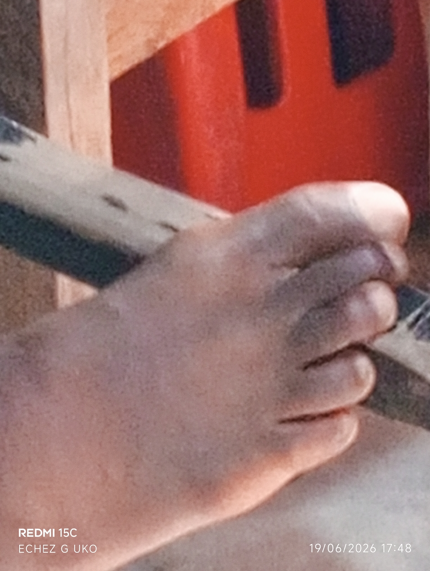 A close-up shot focuses on a person's right foot, with toes curled and resting on a textured surface. The skin tone is medium brown, with visible wrinkles and creases on the toes. A patterned object, possibly a strap or fabric, runs diagonally across the image behind the foot. In the background, out of focus, are elements of what appears to be outdoor furniture, with sections of warm-toned wood and a bright red object. The bottom left corner displays text: "REDMI 15C ECHEZ G UKO". The bottom right corner shows a date and time: "19/06/2026 17:48". The image appears to be taken in Jalingo, Nigeria.