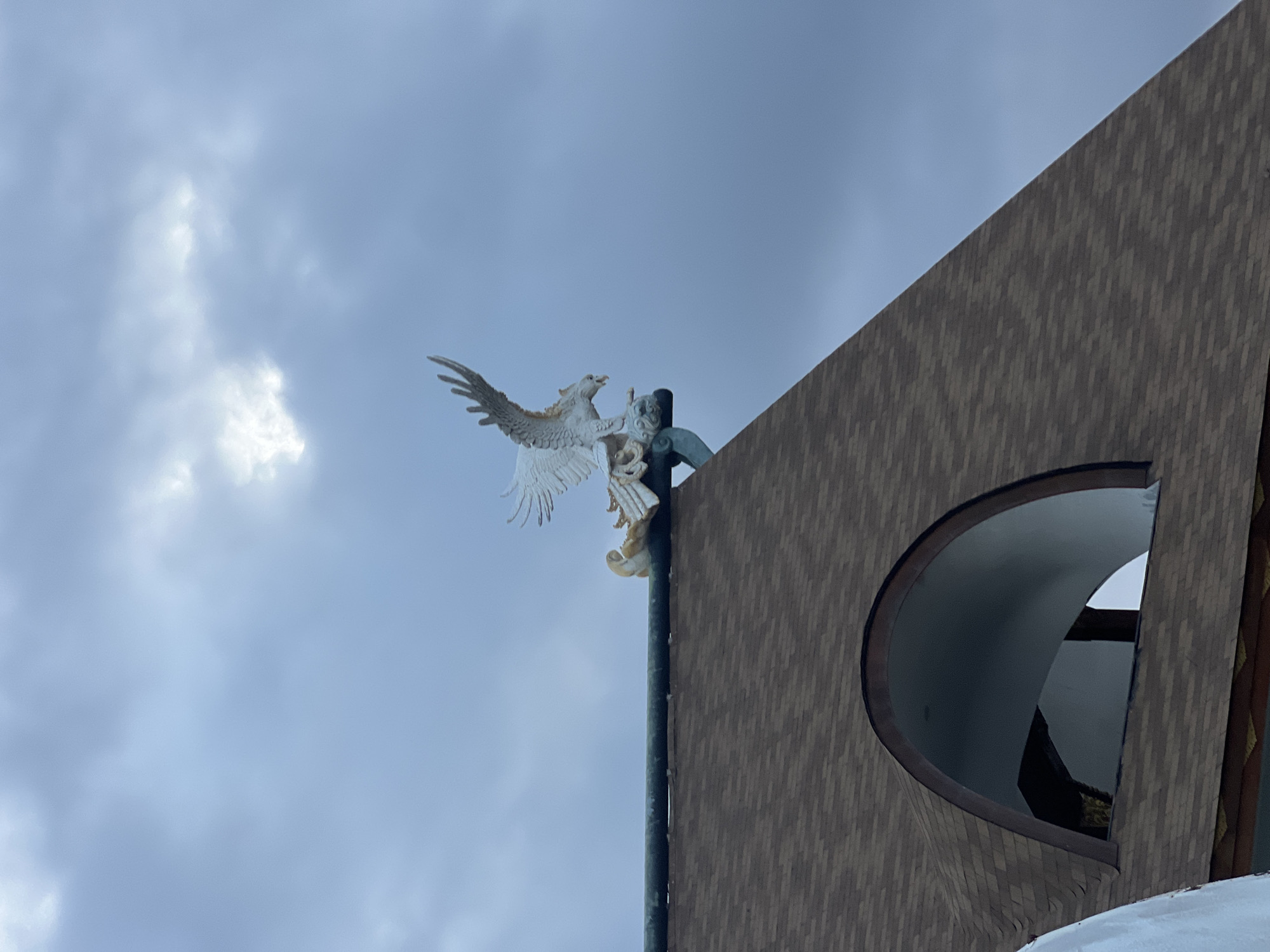 An upward-looking photograph captures a white, winged sculpture set against an overcast sky, with a portion of a building on the right. The central subject is a detailed sculpture of a mythical creature, possibly a phoenix or a dragon, with its large, textured wings spread wide. It is depicted perching or ascending on a dark, vertical pole. Below the primary winged figure, another smaller, human-like or angelic figure is incorporated into the sculpture, appearing to hold onto the pole or the main creature.
The sky is a soft, light blue, filled with scattered white and grey clouds, indicating an overcast daytime setting. On the right side of the frame, a modern building is visible, featuring an exterior covered in small, dark, rectangular tiles or shingles arranged in a diagonal pattern. A distinctive architectural element of the building is a large, deep, semicircular or D-shaped opening, which appears to be a window or alcove, revealing a dark interior. No human activity or visible text is present in the image. The sculpture's design, particularly the winged creature, aligns with mythical motifs found in Indonesian culture, consistent with the provided Denpasar location context.