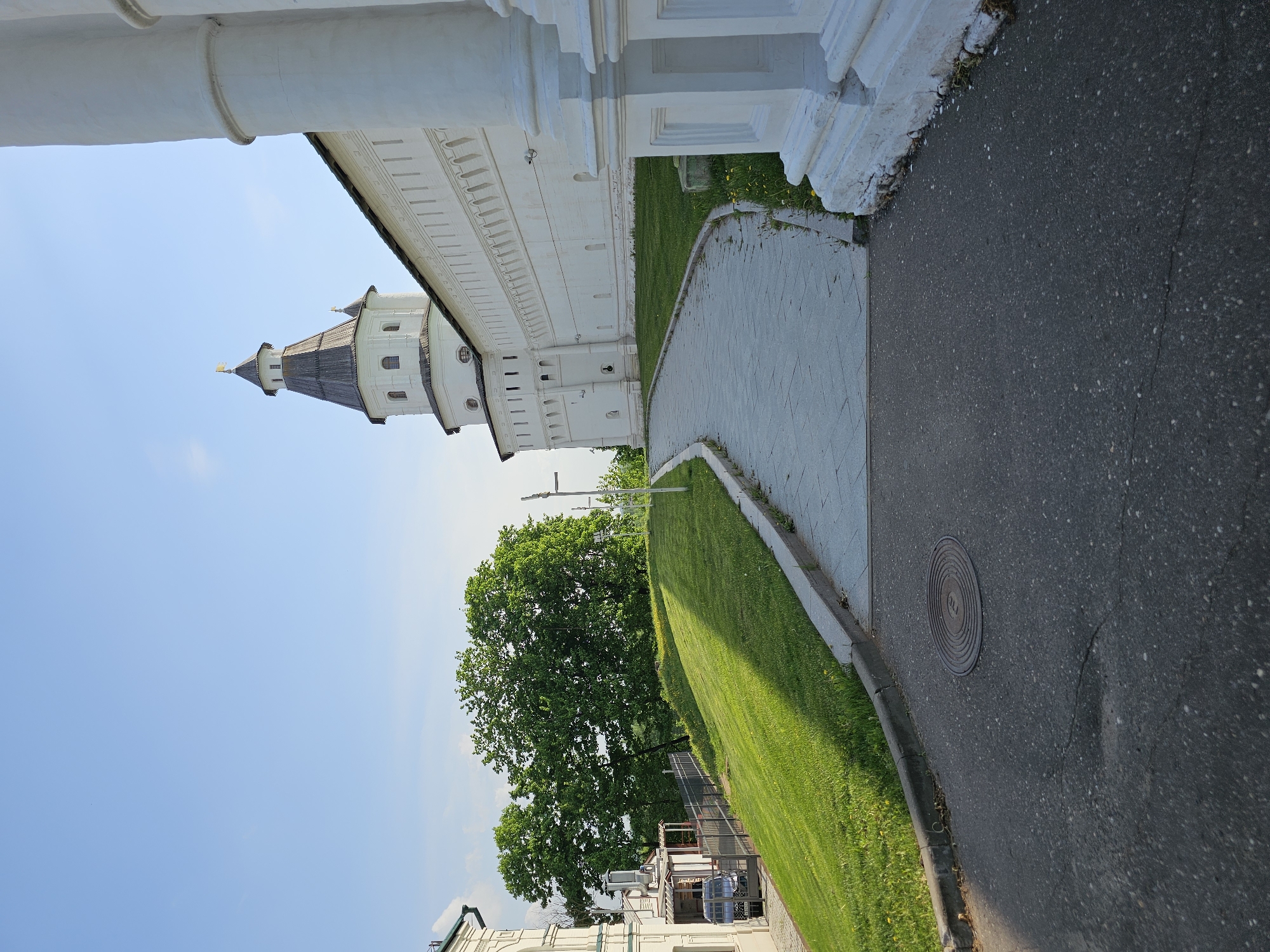 The image shows a view of the New Jerusalem Monastery in Istra, Russia. The viewer is looking at a section of the white-walled monastery with a tower and spire visible against a blue sky. Below the monastery wall is a grassy slope, a paved walkway, and a paved road. The manhole cover is in the pavement. A green tree is visible on the slope.