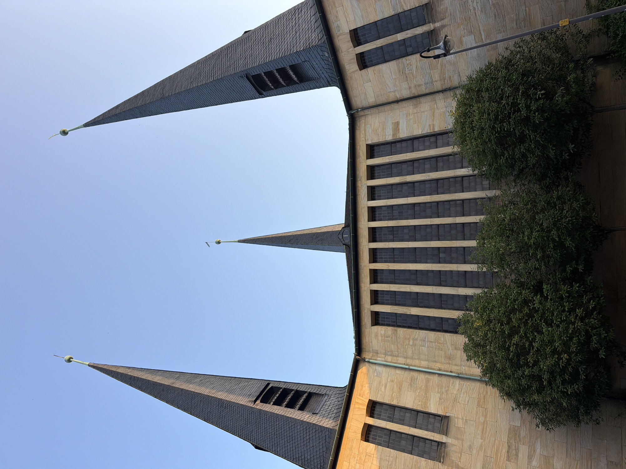 The image shows an upward view of the exterior of a church in Bayreuth, Germany. The church has a modern architectural style, characterized by a facade made of light-colored stone blocks and tall, narrow windows with dark panes. Three prominent spires rise from the building, two on the sides and one in the center. The spires are covered in dark grey slate tiles and topped with globes and crosses. The spires on the sides have rectangular openings, suggesting they may house bells. Lush green trees are visible in the foreground, partially obscuring the base of the church. The sky is clear and blue, indicating a daytime scene. A street lamp is visible on the right side of the image. The overall impression is one of grandeur and a blend of historical and contemporary design elements.