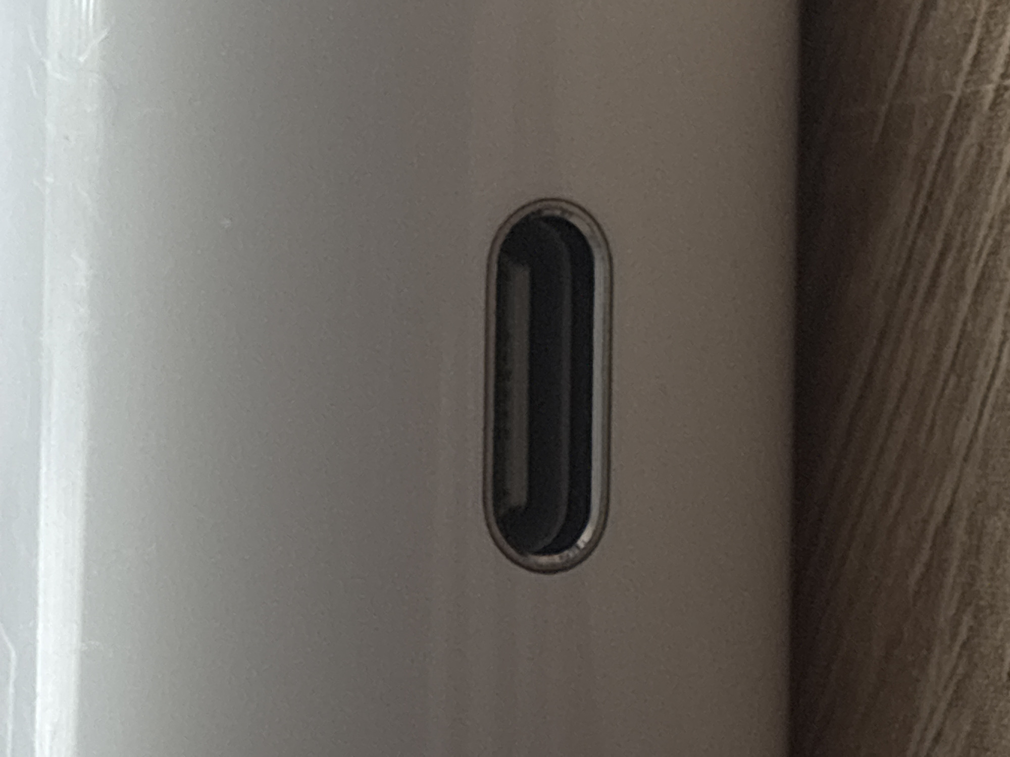 The image shows a close-up, eye-level view of a USB-C port on a white device. The port is rectangular with rounded corners and appears to be made of metal. The device is resting on a wooden surface with a visible grain pattern. The lighting is soft, and the background is out of focus. There are no people or any other objects visible in the scene. This image is not related to the city of Saint Petersburg, Russia.