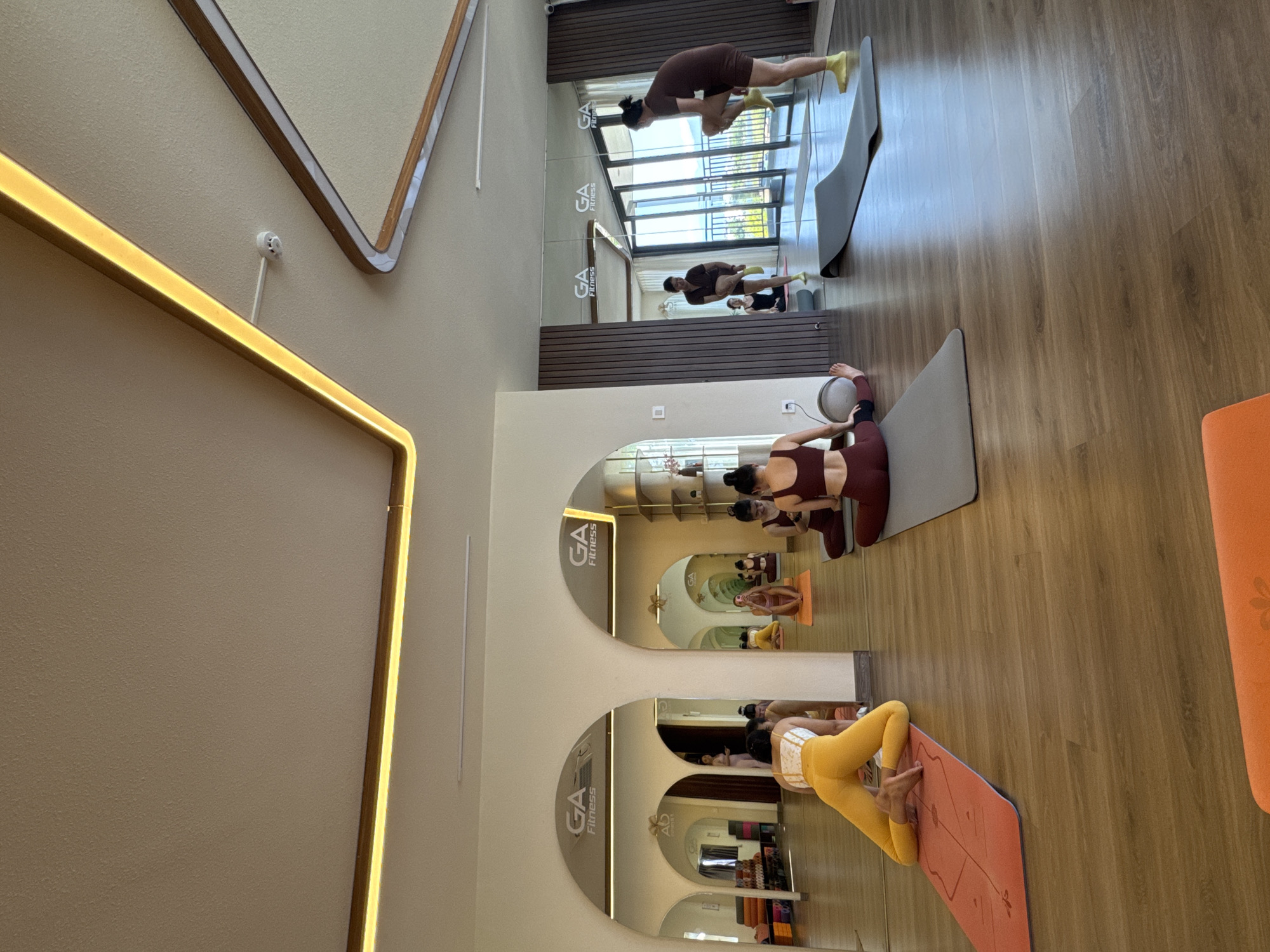 Here is a detailed description of the image:
The image shows the interior of a yoga studio. Several women are visible, performing yoga poses on mats. They wear athletic attire. Mirrors line the walls of the studio, reflecting the women and creating the illusion of a larger space. The studio has a light and airy feel. The floors are wooden, and the walls are painted in light neutral tones.
The studio appears to be well-lit with natural and artificial lighting. The large windows offer a view of the outside. The location is a yoga studio. The city and place is unknown.