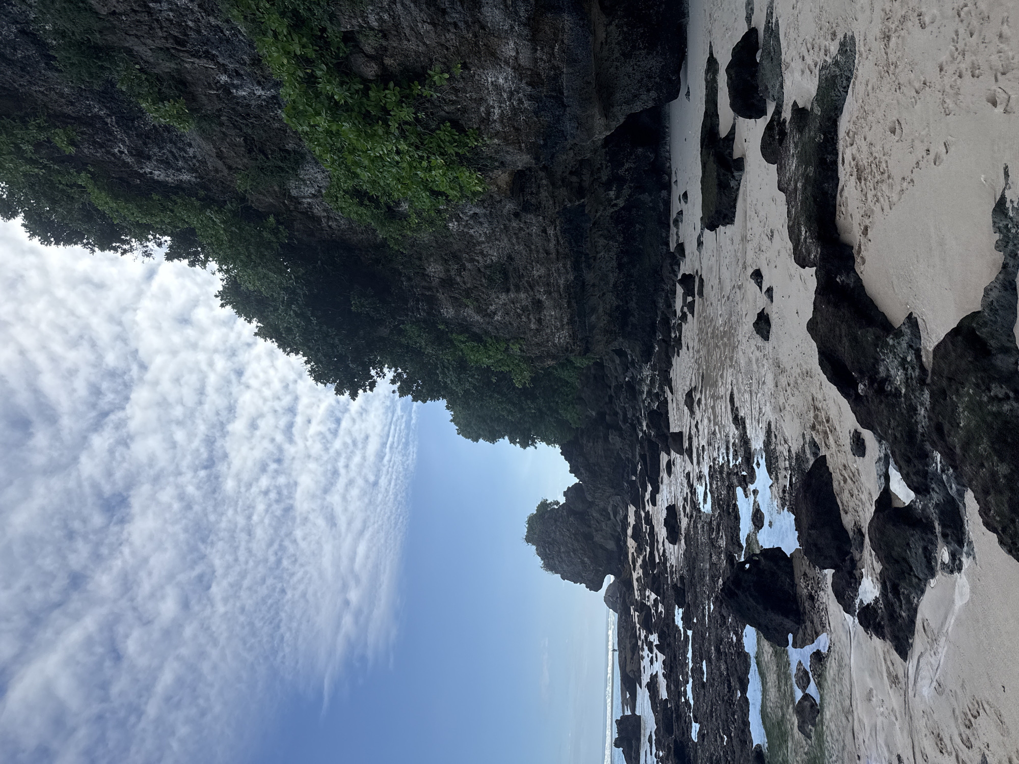 The image shows a view of a coastline in Kuta Selatan, Indonesia. A rocky cliff face covered in vegetation occupies the top left of the frame. The sky is blue with scattered clouds. Below the cliff is a beach with a mix of white sand and dark rocks. Water is visible amongst the rocks near the shoreline. In the distance, a person stands on the beach.