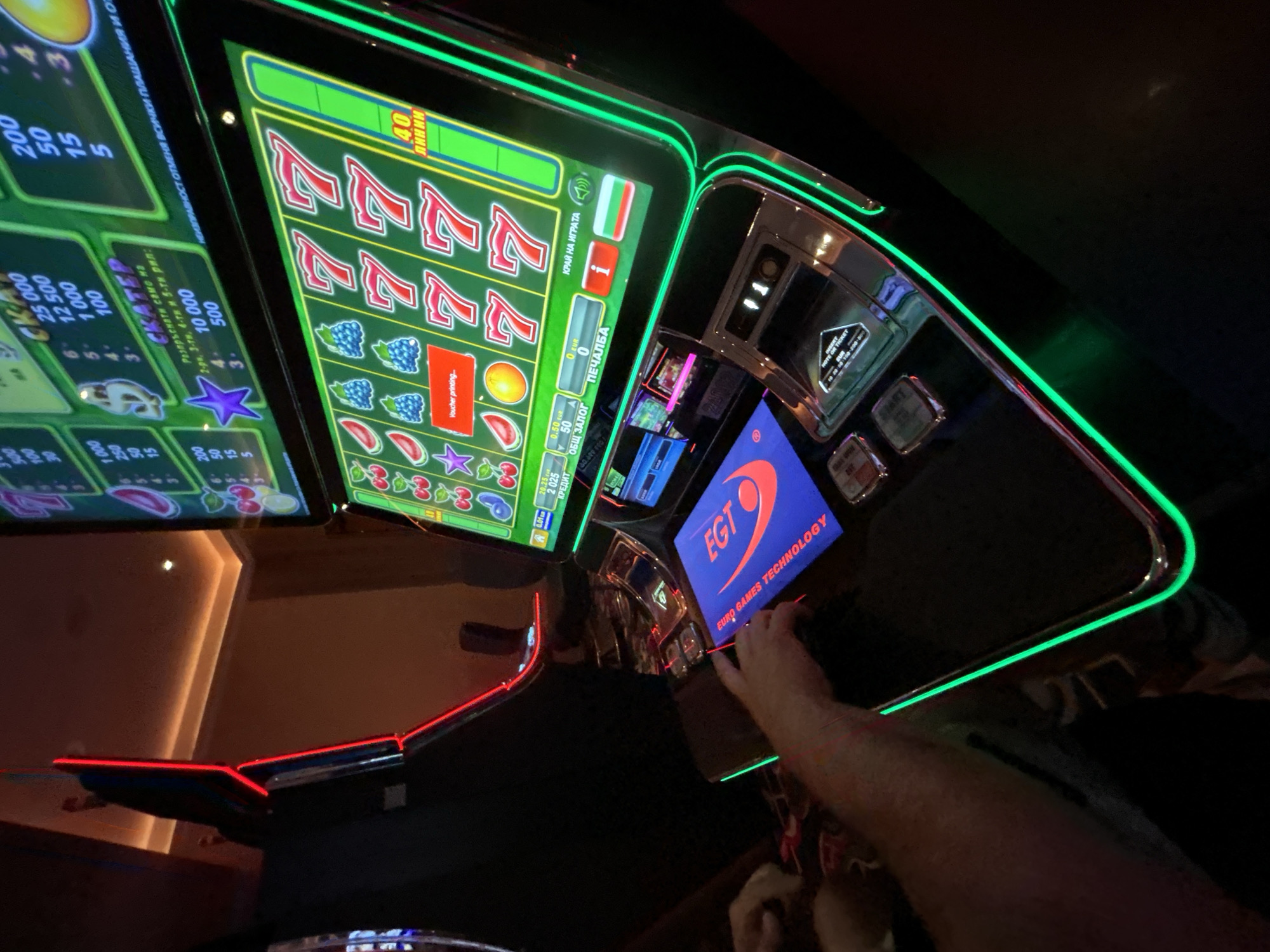 The image shows a close-up view of a slot machine in a casino. The slot machine has a vibrant green display with various symbols and numbers, including cherries, berries, watermelons, oranges, stars, and the number '7'. The machine is lit by bright green neon lights around its edges. In the foreground, a blue touchscreen panel displays the EGT logo and the text "EURO GAMES TECHNOLOGY". A person's hands are visible interacting with the machine, suggesting they are playing. The background shows a dimly lit casino environment with other slot machines and ceiling lighting. The image is taken from a medium to low angle. It is not possible to definitively determine the location within the city of Vratsa, Bulgaria, from the visual information provided.