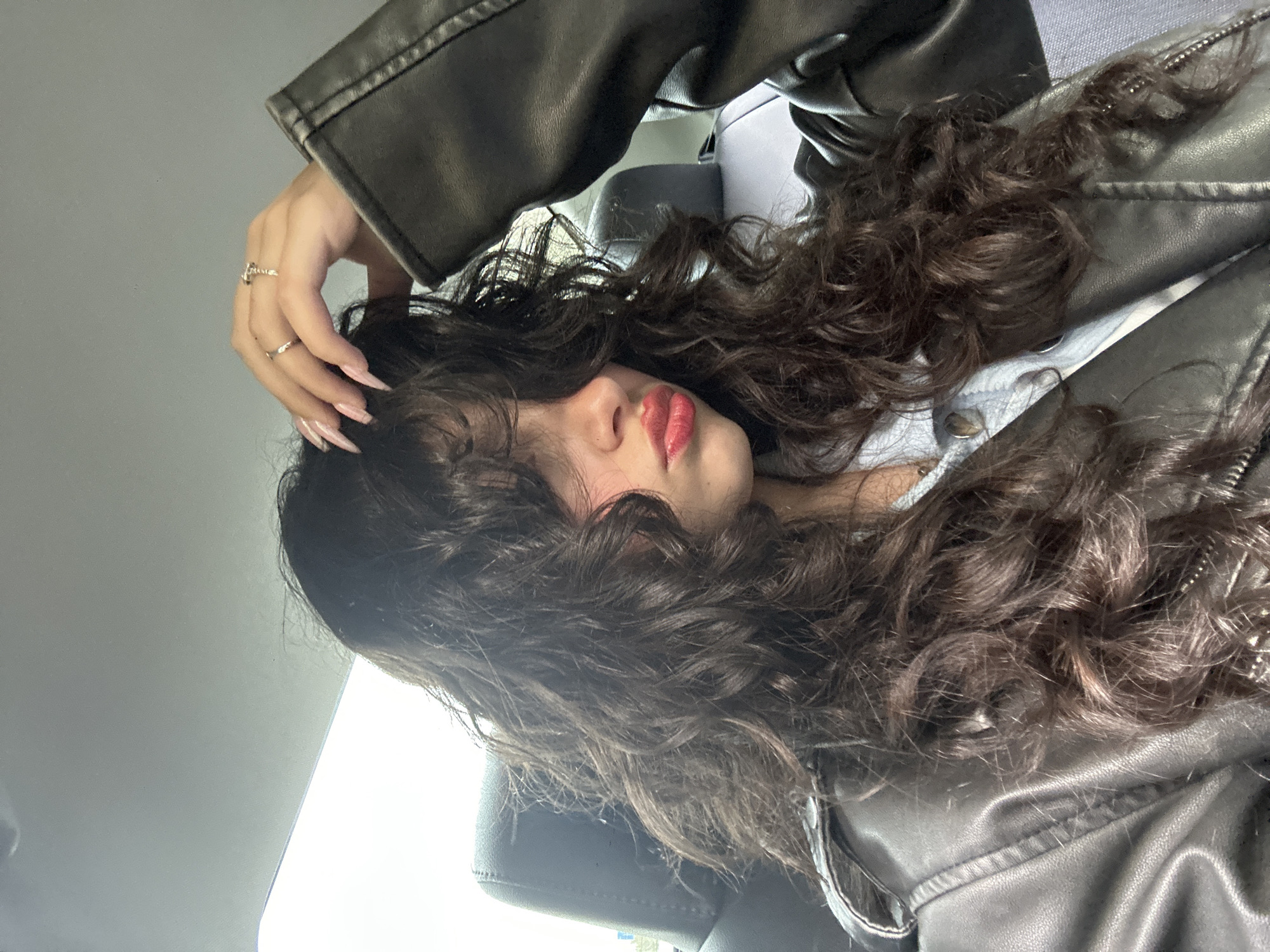 A dark-haired person with long, wavy hair and red lipstick is reclined, possibly in a car seat, wearing a black leather jacket and a light-colored top. The person's left hand, adorned with rings and long pink nails, rests on their head. The background includes a car interior and potentially an exterior view of Yerevan, Armenia, but specific landmarks or distinguishing features are not visible, making positive confirmation of the location impossible.