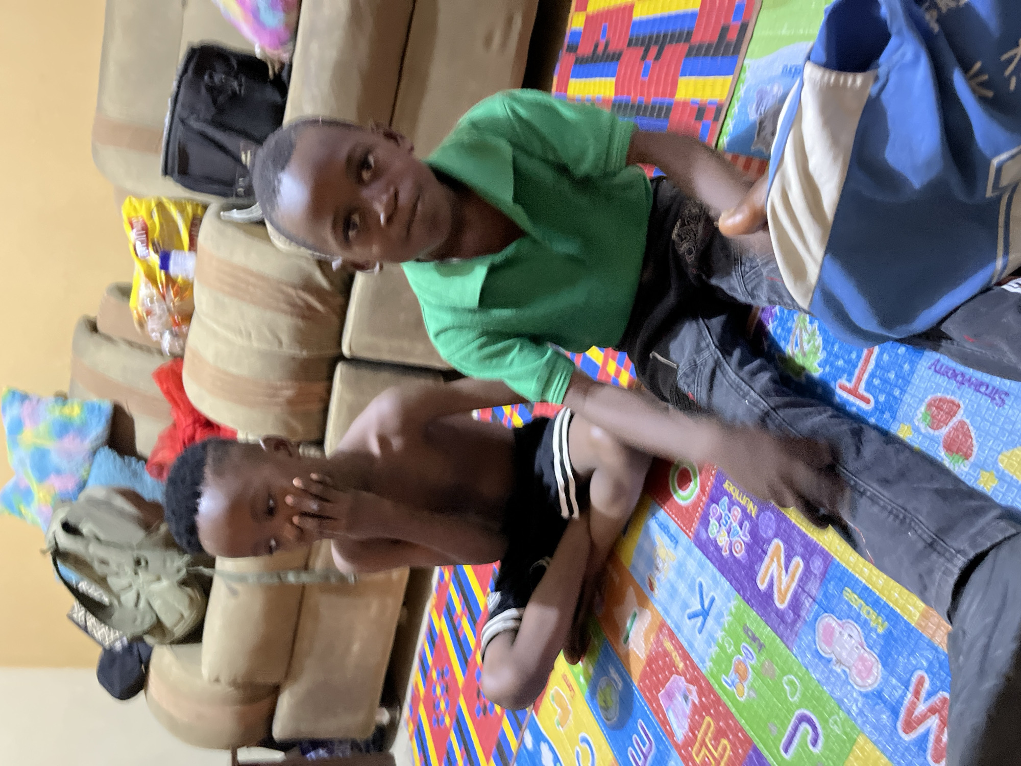 Two young Black children are indoors, sitting on a colorful ABC alphabet play mat. One child wears a green polo shirt and dark jeans, while the other is shirtless with black and white striped shorts. The play mat is on a floor and extends onto a beige couch. In the background, there is a backpack and other items on the couch. The location is Madalla, Nigeria.