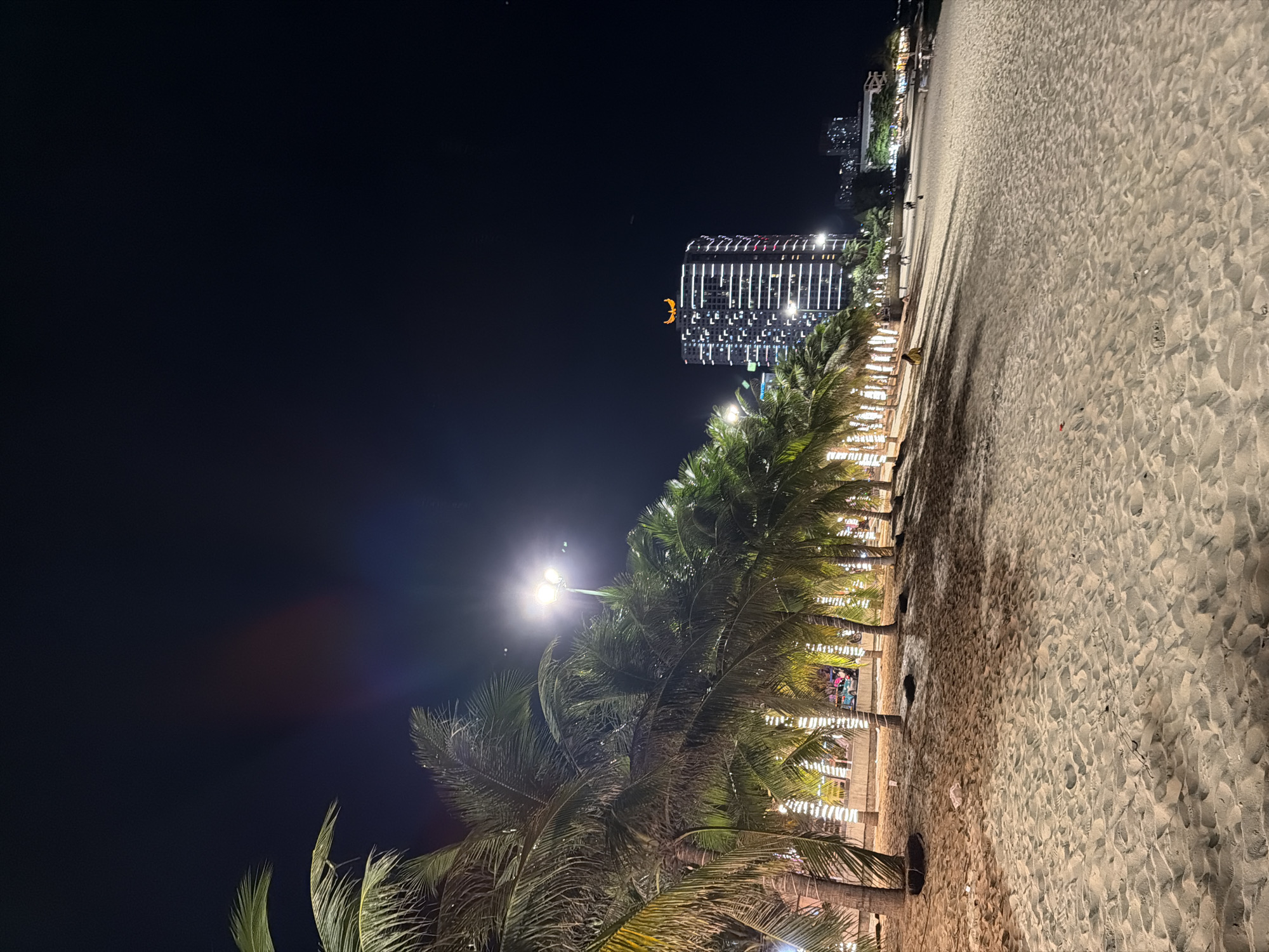 The image captures a nighttime scene of a sandy beach in Nha Trang, Vietnam, with palm trees lining the shore. The trees are illuminated by string lights wrapped around their trunks, creating a festive atmosphere. In the background, a tall, modern building with glowing windows stands out, topped with a golden, winged emblem. Other buildings and lights are visible in the distance. The sandy beach shows footprints and scattered debris, suggesting it is a public space with recent activity. The sky is dark, indicating it is late evening or night. A few people can be seen walking on the beach and near the building. The overall scene depicts a vibrant urban beach at night, likely a popular tourist spot.
