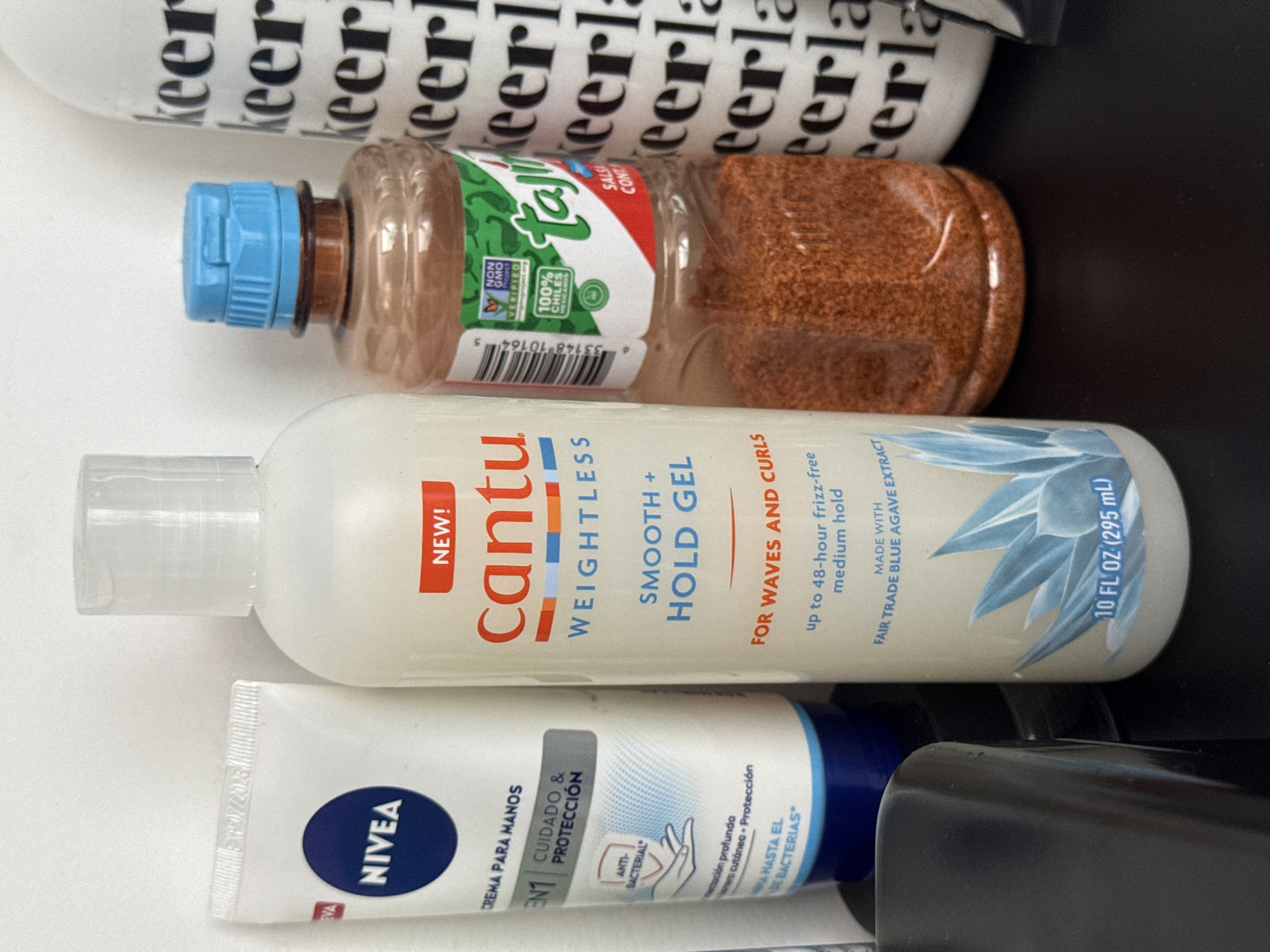 This image shows an assortment of personal care and food products. The items are, from top to bottom:
1. A bottle with "Keerl" printed repeatedly on it.
2. A plastic bottle of Tajin seasoning with a blue lid.
3. A bottle of Cantu Weightless Smooth + Hold Gel for waves and curls.
4. A tube of Nivea hand cream, labeled in Spanish.
The location is not discernable from the image, and there's no basis for stating it's San Bernardino Tlaxcalancingo, Mexico. No people or interactions are visible. The environment is a neutral, light-colored surface.