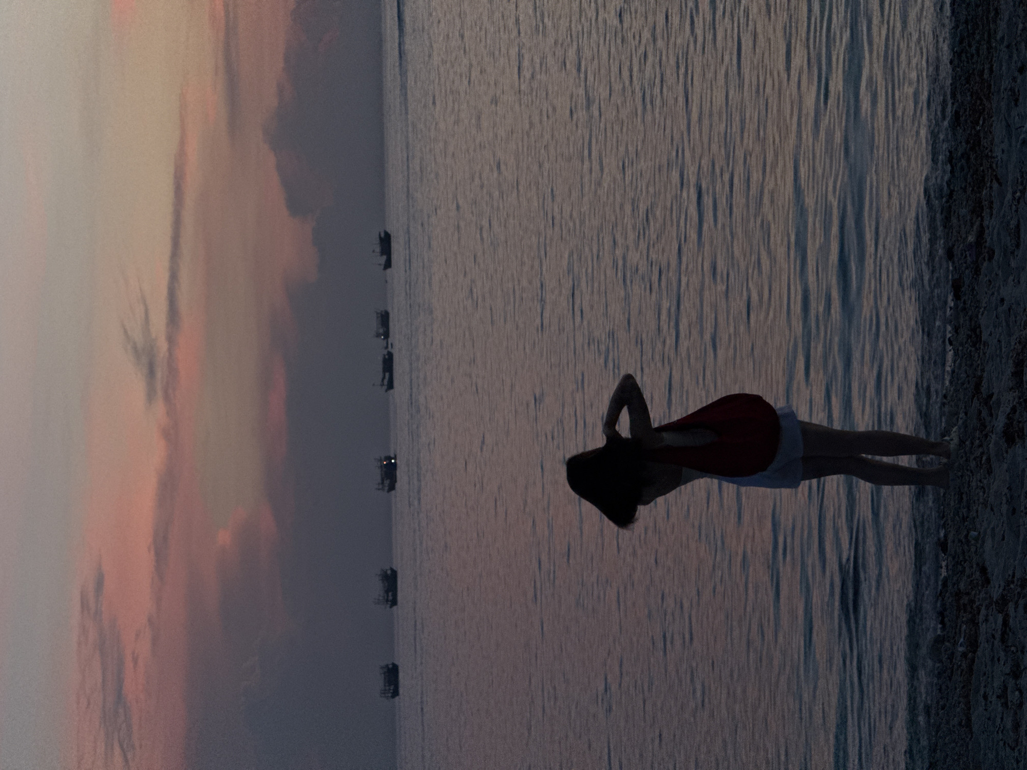 The image depicts a silhouette of a person standing on a beach at dusk. The person, facing away from the camera, has dark hair and is wearing light-colored shorts and a tank top. A red bag is slung over their shoulder. Their arms are raised, with hands near the back of their head, possibly adjusting their hair or looking through a viewfinder. The ocean stretches out before them, with gentle waves lapping at the shore. In the distance, several small fishing boats are visible on the horizon. The sky is painted with soft hues of pink and purple, indicative of a sunset. The beach itself appears to be composed of sand and pebbles. The overall atmosphere is tranquil and serene. This scene is characteristic of the coastal areas of Lý Sơn Island, a special administrative region in Vietnam, known for its fishing communities and beautiful sunset views.
