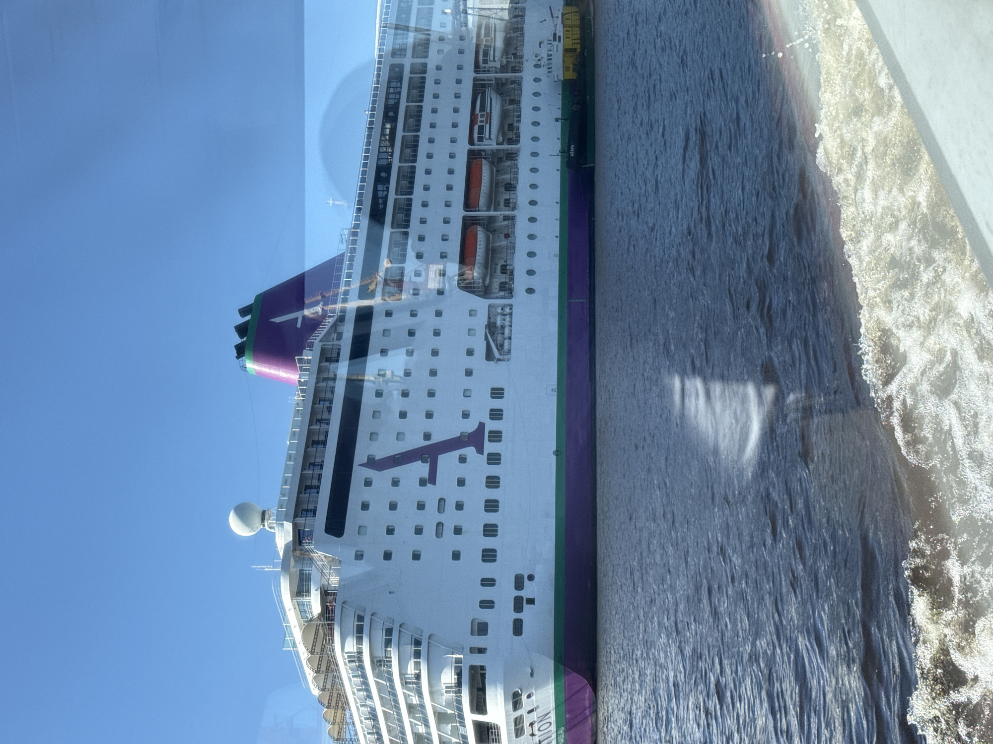 A large white cruise ship with purple and green accents is docked in the Ventspils Port, Latvia. The ship is prominently displayed against a clear blue sky. The word "STENA" is visible on the hull of the ship. A smaller boat with the name "UZAVA" on its side is moored next to the cruise ship. The water in the foreground is choppy, suggesting movement from a boat. The overall scene depicts a busy port environment on a sunny day.