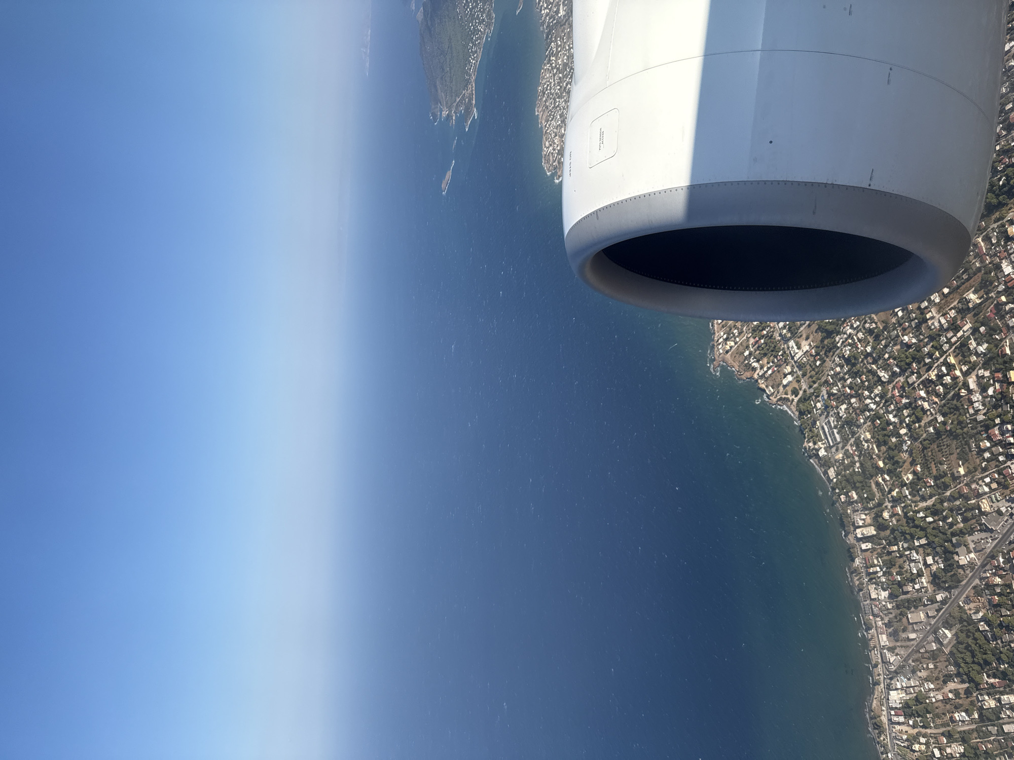 An aerial, slightly elevated, medium shot captures a view from an airplane window, featuring the ocean and a coastal city. In the foreground, a portion of the airplane's engine cowling occupies the right side of the frame. The cowling is white with a dark gray inlet. The ocean, a deep blue, stretches across the majority of the image, meeting a shoreline dotted with buildings and vegetation. The city appears to be built on a gentle slope leading down to the water. The sky is a clear, bright blue, with no visible clouds. The scene appears to be in Artemida Municipal Unit, Greece, given the distinctive coastline and urban development visible.