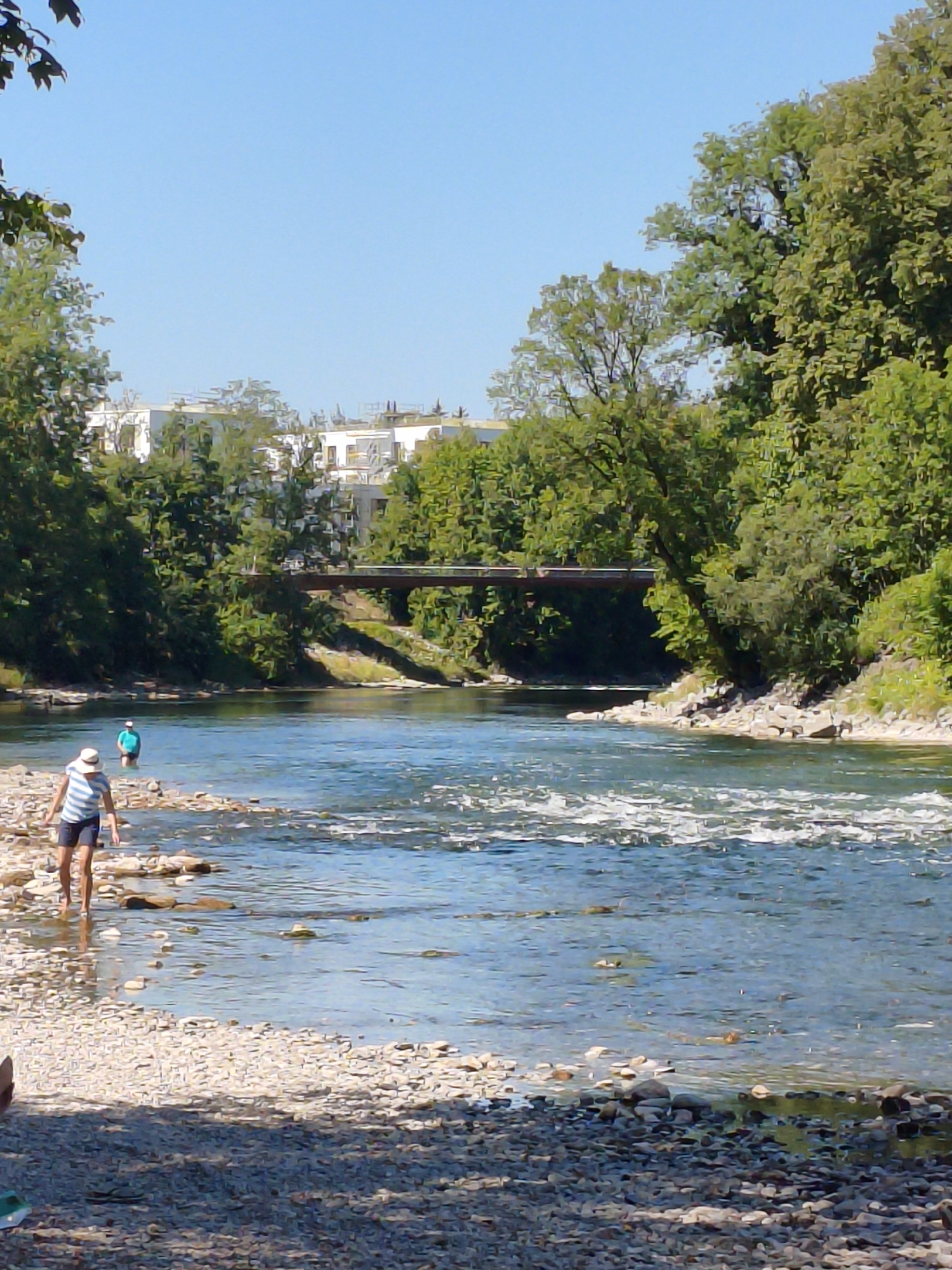 The image shows a sunny day scene at the Lech river in Landsberg am Lech, Germany. In the foreground, a person wearing a white and blue striped shirt and dark shorts stands on a rocky shore, facing the water. Further out in the river, another person in a blue shirt is visible. The river has clear, blue water with ripples and some submerged rocks. Trees line the banks of the river, and in the background, a bridge crosses the water, with buildings and more trees visible beyond the bridge. The sky is clear and blue.