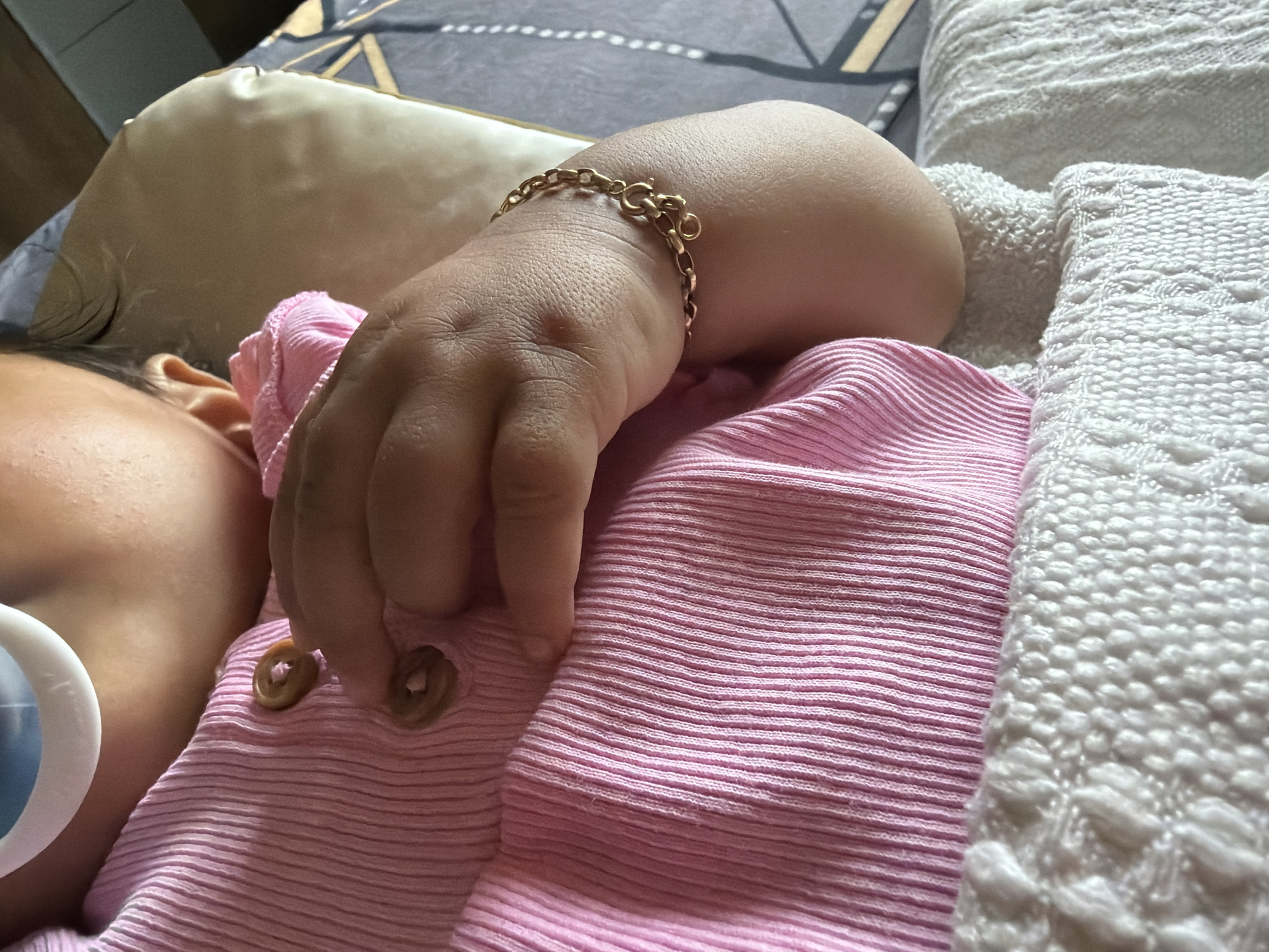 A close-up shot shows a baby's hand resting on their chest, wearing a pink ribbed outfit and a gold chain bracelet. The baby is also wearing a pacifier. The background appears to be a bed with a textured white blanket and a patterned sheet. The image is taken indoors, likely in a home environment. There is no discernible information within the image to indicate the location of Razhevo Konare, Bulgaria.