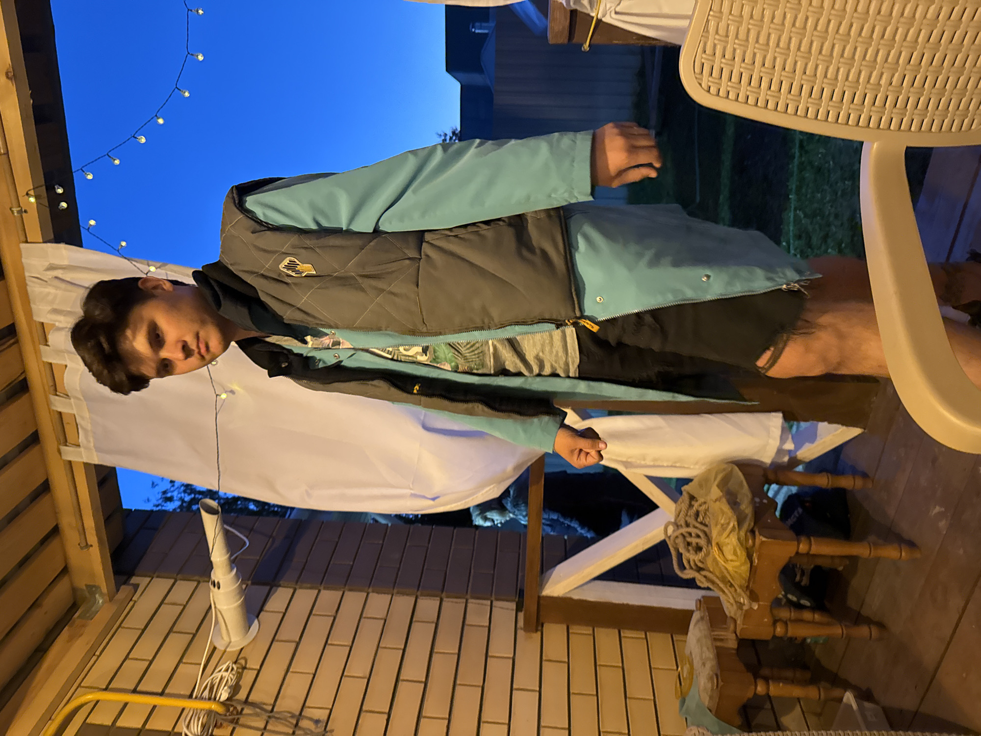 A young male stands on a wooden porch at dusk. He is wearing a grey puffer vest over a turquoise jacket and shorts. He is looking away from the camera. In the background, a white curtain hangs, and string lights are visible against the dark blue sky. To the left of the male, a brick wall is visible, with a yellow pipe attached to it. There are two wooden stools with items on them, and a chair is in the foreground. The overall scene suggests an outdoor gathering or casual setting.