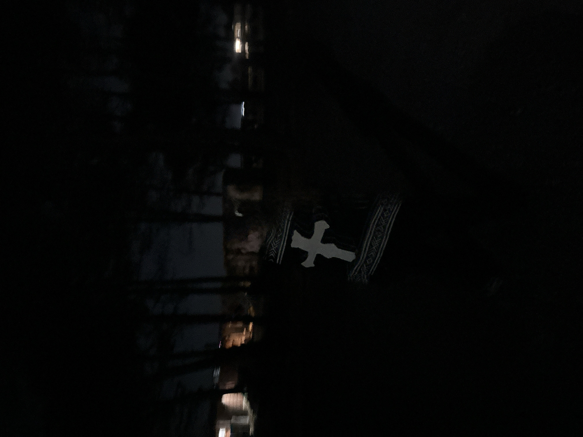 A dark, nighttime image appears to be taken in Donji Štoj, Montenegro. In the background, faint reflections of lights suggest buildings or streetlights. The central subject is a person wearing a black garment with a large, white cross emblem visible on the front. The person's face is obscured by shadows.