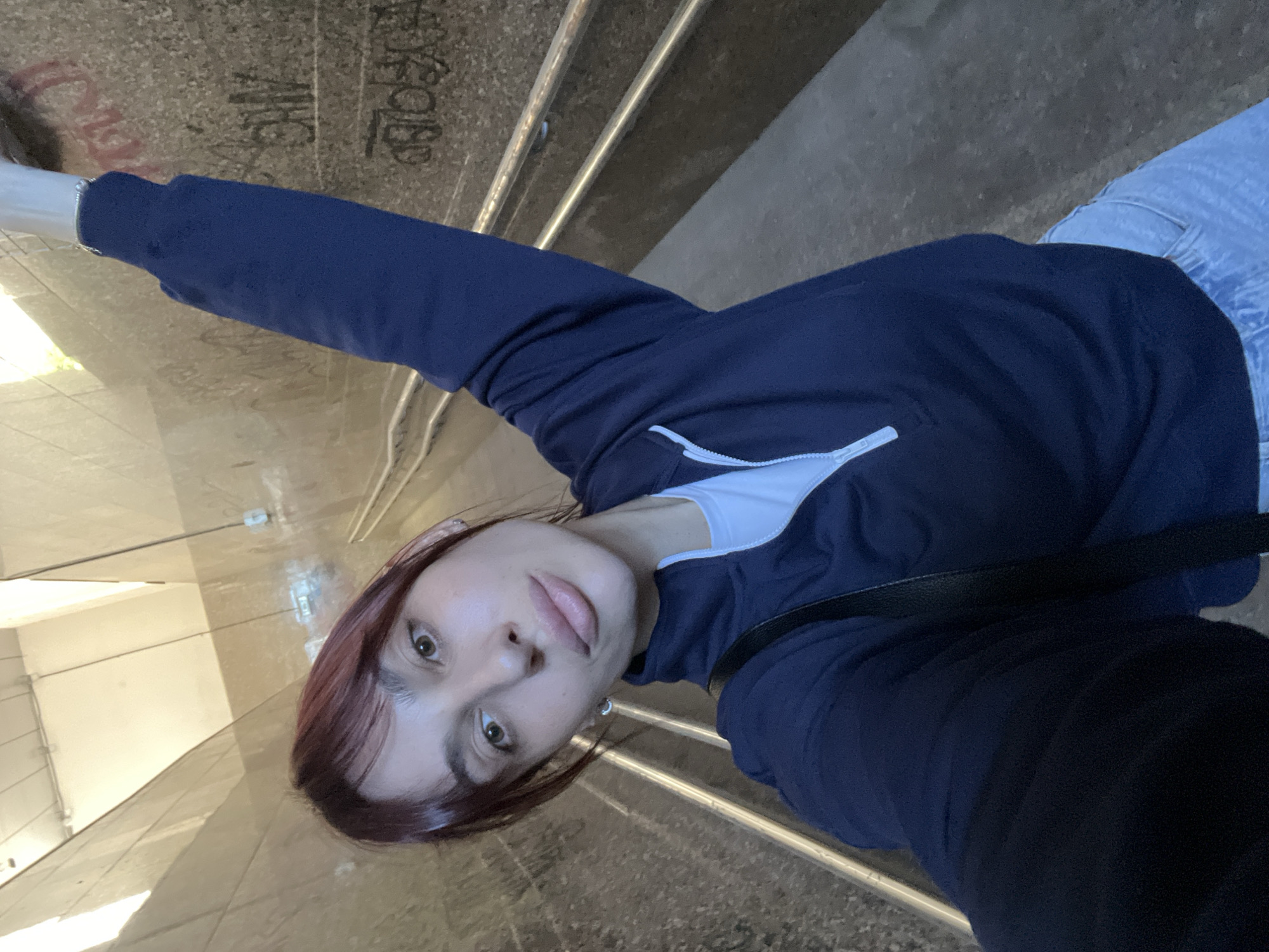 A young woman with red hair and light skin is taking a selfie in a graffiti-covered underground passage in Nizhny Novgorod, Russia. She is wearing a dark blue sweatshirt and light blue jeans, and her right arm is raised. The passage has concrete walls, metal railings, and overhead lighting. The lighting in the passage is artificial and appears to be fluorescent.