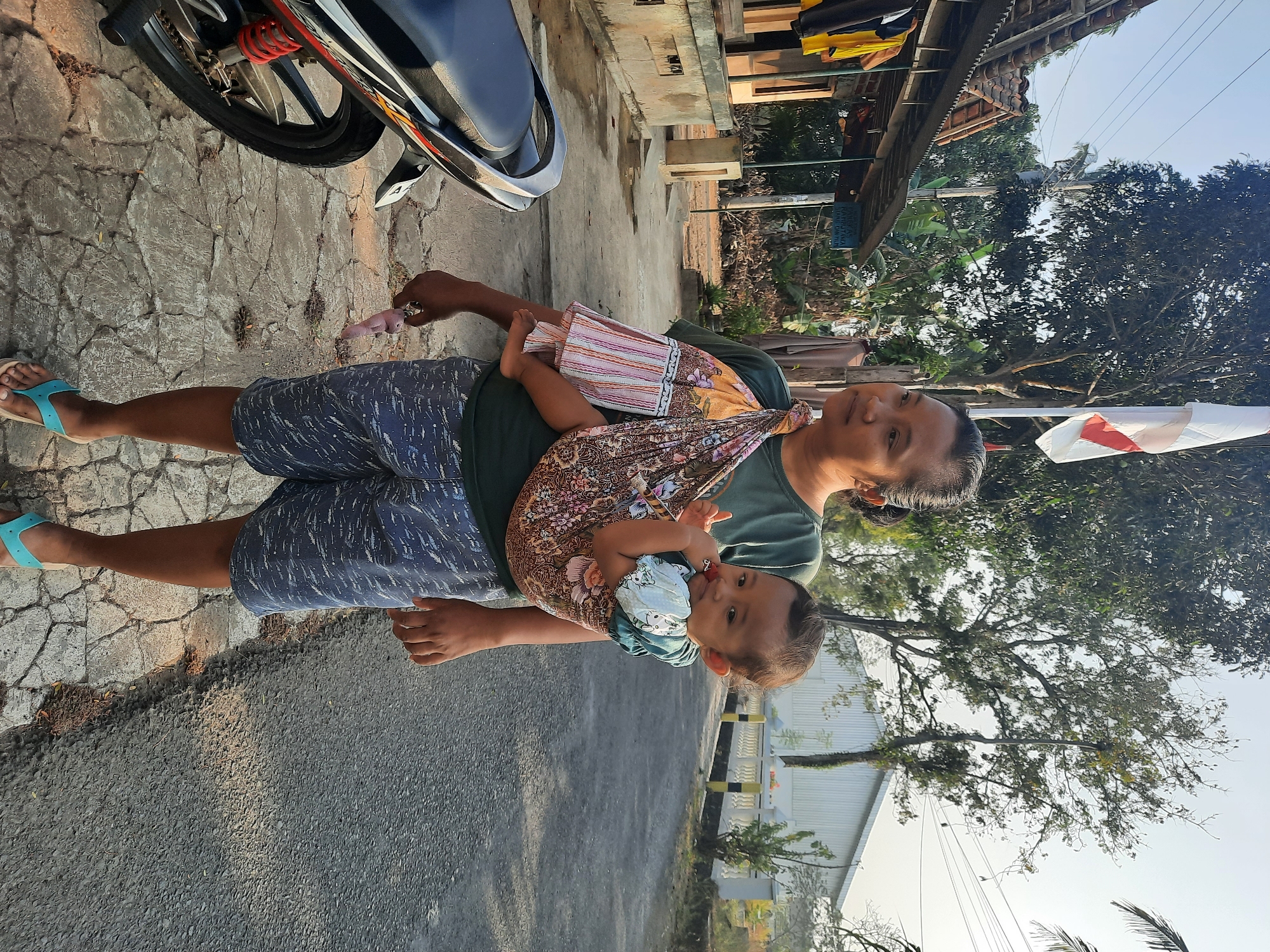 A woman is carrying a baby on her back in a sling in the village of Andong, Indonesia. The woman is wearing a dark green t-shirt and blue patterned shorts. The baby is wearing a light blue shirt and is looking at the camera. They are standing on a cobblestone path next to a road. A motorcycle is parked on the left side of the image. In the background, there are trees, a building with a white roof, and a flag on a flagpole. The sky is clear and the sun is shining.