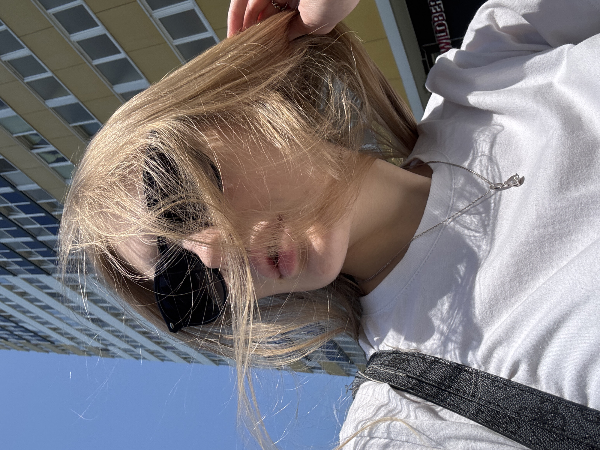 A blonde woman wearing sunglasses and a white t-shirt stands outside in Perm, Russia. She is partially visible in the frame, from the neck up, and is touching her hair with her right hand. The building at Permyakova street 1, which has a grid of windows, is visible in the background. A silver necklace is visible on the woman's neck. The sky is blue.