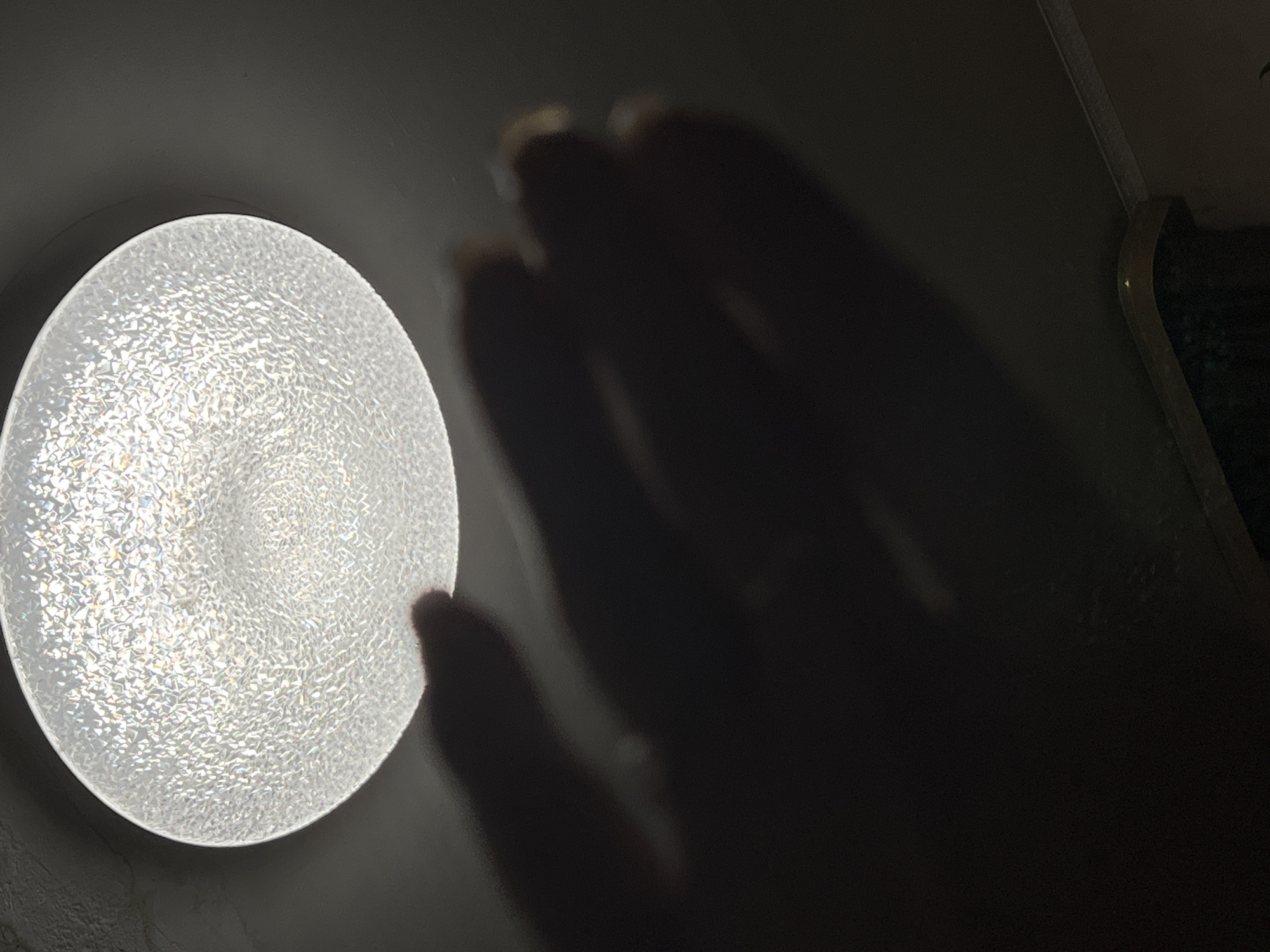 The image shows a close-up view of a ceiling light fixture, with a hand casting a shadow on it. The light fixture is round and appears to be made of a textured, translucent material, possibly glass or plastic, that diffuses the light creating a speckled effect. The hand is in the foreground, silhouetted against the bright light of the fixture, obscuring a portion of it. The fingers are spread, and the shadow they cast is large and somewhat indistinct due to the proximity to the light source. The background is dark, indicating that the surrounding room is dimly lit. The image was captured in Yekaterinburg, Russia.