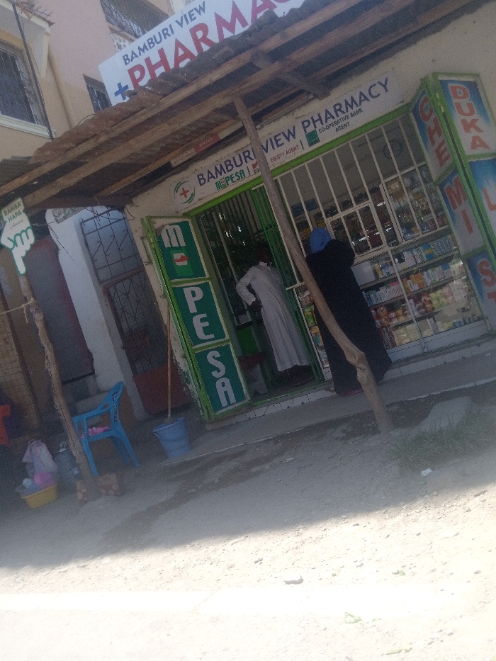 This image captures the exterior of the "BAMBURI VIEW PHARMACY" and M-PESA agent in Mtopanga ward, Kenya. The storefront features a white facade with green trim, displaying a primary sign for "BAMBURI VIEW PHARMACY" with a green cross logo. Additional signage indicates it functions as a "CO-OPERATIVE BANK AGENT" and "EQUITY AGENT." Vertical green panels on either side of the entrance prominently display "M-PESA." A partially visible sign on the far right reads "DUKA CHEMIST."
An individual in a light-colored robe is positioned inside the pharmacy's entrance, facing away from the camera. Another individual, wearing a dark abaya and a light blue hijab, stands just outside the entrance, facing into the pharmacy. Visible through the barred entrance are interior shelves stocked with various products.
A makeshift overhang constructed from wooden beams and corrugated material extends over the storefront. The ground in front of the establishment is unpaved and dusty. To the left, a blue plastic chair, a yellow basin containing items under a clear plastic cover, and a blue bucket are present. A small white sign featuring a green hand symbol and the text "BAMBA HAPA" is attached to a wooden post on the far left. Sparse patches of green grass are visible on the ground to the right. The scene is illuminated by natural daylight.