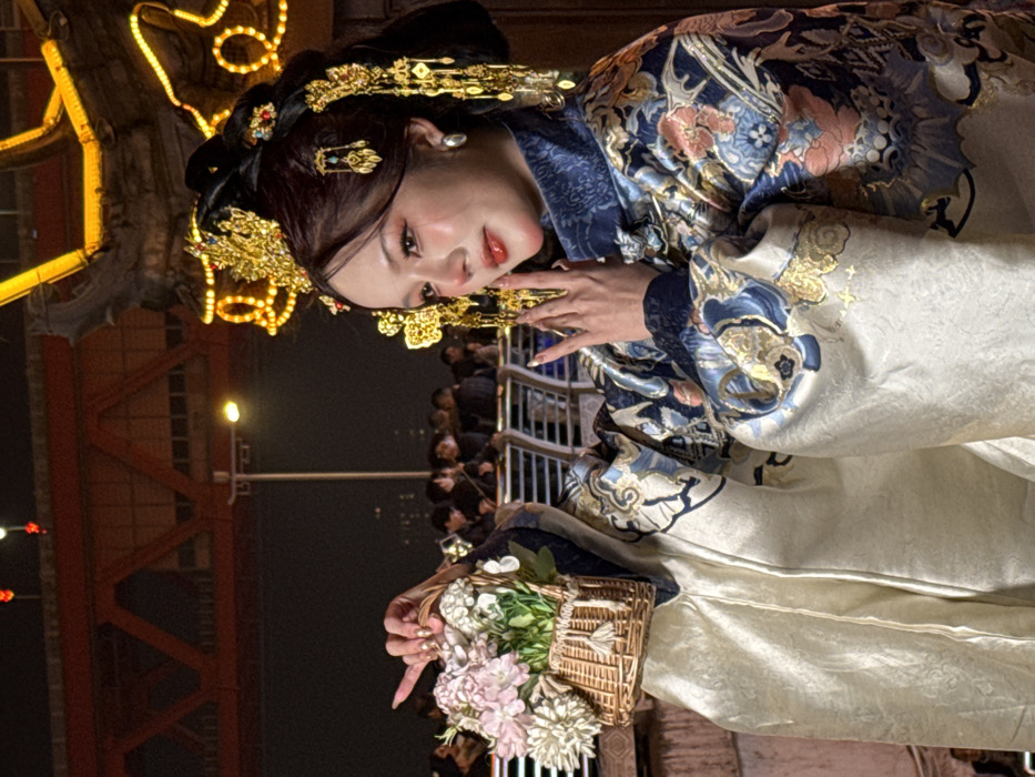 The image depicts a woman in ornate traditional Chinese attire, posing at night in Yuzhong District, China. She wears a blue and gold patterned robe adorned with intricate floral and cloud designs, over a cream-colored inner garment. A lavish golden headdress featuring jewels and dangling ornaments is prominently displayed on her dark hair. Her left hand holds a small woven wicker basket filled with artificial pink and white flowers, while her right hand is raised to her chest in a stylized gesture. Her fingernails are painted a metallic gold. She looks slightly to her right with a gentle expression.
In the background, a metal railing separates the subject from a blurry crowd of people. Further back, a large red steel truss bridge is visible on the left, with subtle red lights indicating its structure. To the right, a large, ornate structure, possibly an archway or building element, is outlined with golden string lights. The scene is illuminated by artificial night lighting, including a bright street lamp positioned behind the woman.