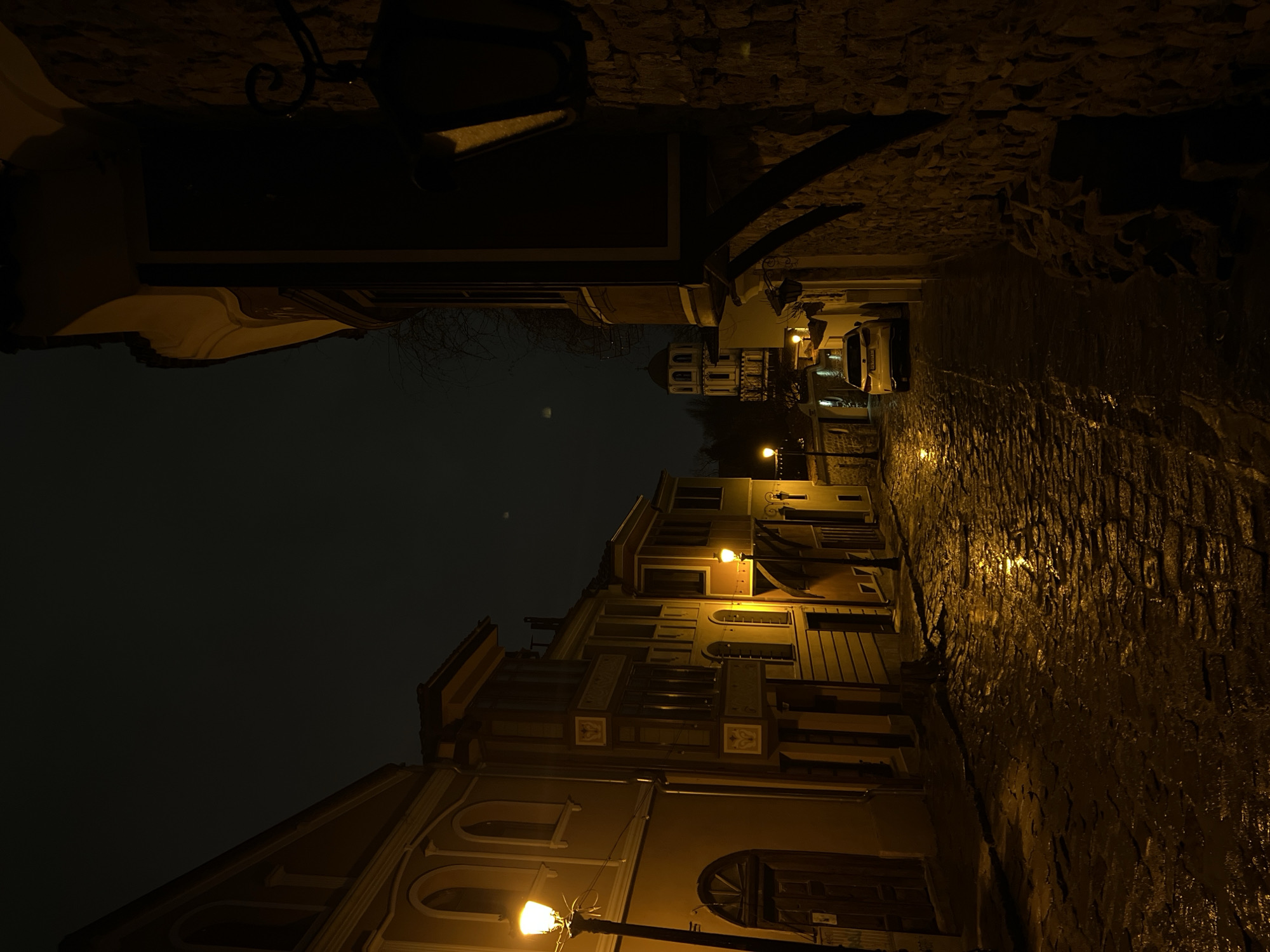 This image depicts a narrow, wet cobblestone street at night within the Old Town of Plovdiv, Bulgaria. The street slopes gently downwards and is illuminated by several warm yellow streetlights, whose reflections are visible on the damp pavement.
On the left, a row of traditional Plovdiv Revival-style houses features multi-story facades, some with ornate bay windows, arched ground-floor entrances, and decorative cornices. Colors appear as muted yellows, oranges, and browns under the artificial light. On the immediate right, a dark stone wall forms the foreground, partially obscuring a building with an overhanging upper floor.
Further down the street, a white passenger car is parked on the left. In the background, a distinctive church tower with multiple domed sections, likely the Saints Constantine and Helena Church, is visible against a dark, overcast sky. Two faint, hazy light sources are barely discernible in the upper sky. No individuals are present in the scene.