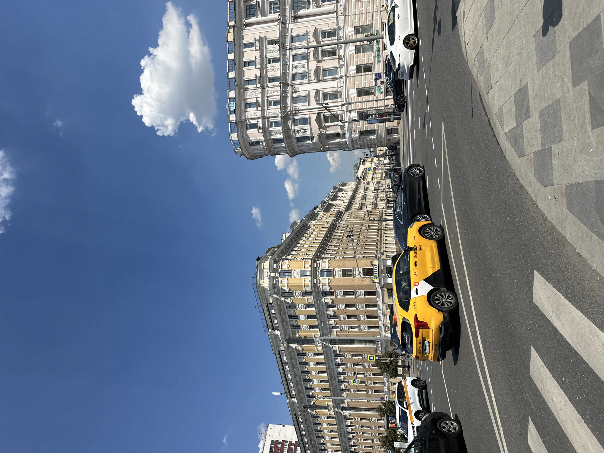 This is a medium-shot, eye-level, outdoor, daytime photograph taken from a street perspective in Moscow, Russia. The scene depicts a busy intersection with several cars, including a bright yellow Yandex taxi in the foreground. The taxi has the number "166" visible on its roof. In the background, ornate, light-colored apartment buildings with multiple windows line the street. The sky is a clear, vibrant blue with a few white cumulus clouds. The buildings exhibit classical architectural elements with decorative moldings and cornices. There are trees and streetlights visible along the sidewalks. The road markings are clearly defined, indicating lanes and a pedestrian crossing. The overall impression is of a sunny day in an urban environment with significant architectural character.