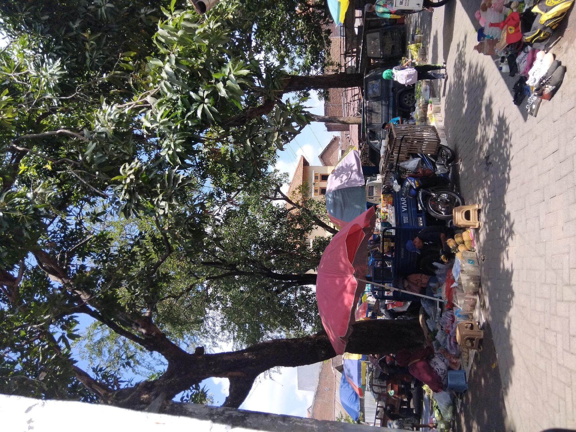 The image depicts a bustling outdoor market scene, likely in the city of Kepuh Kiriman, Indonesia. The foreground is a paved walkway with scattered items for sale, including footwear, bags, and various goods packaged in plastic. Several small stools are also present. A blue pickup truck with the brand name "VIAR" is parked centrally, serving as a vendor's stall. A person wearing a green headscarf and a pink backpack is walking away from the truck. To the left of the truck, a person in a dark hijab and a patterned skirt is seated near a pile of goods. A large red umbrella provides shade over some of the market stalls. Behind the truck, several dark-colored vehicles, including an SUV, are parked. Various other market stalls with umbrellas and a variety of goods are visible in the background, along with buildings with terracotta roofs. Large trees with dense foliage dominate the upper portion of the image, casting dappled shadows on the ground. The sky is blue with scattered white clouds.