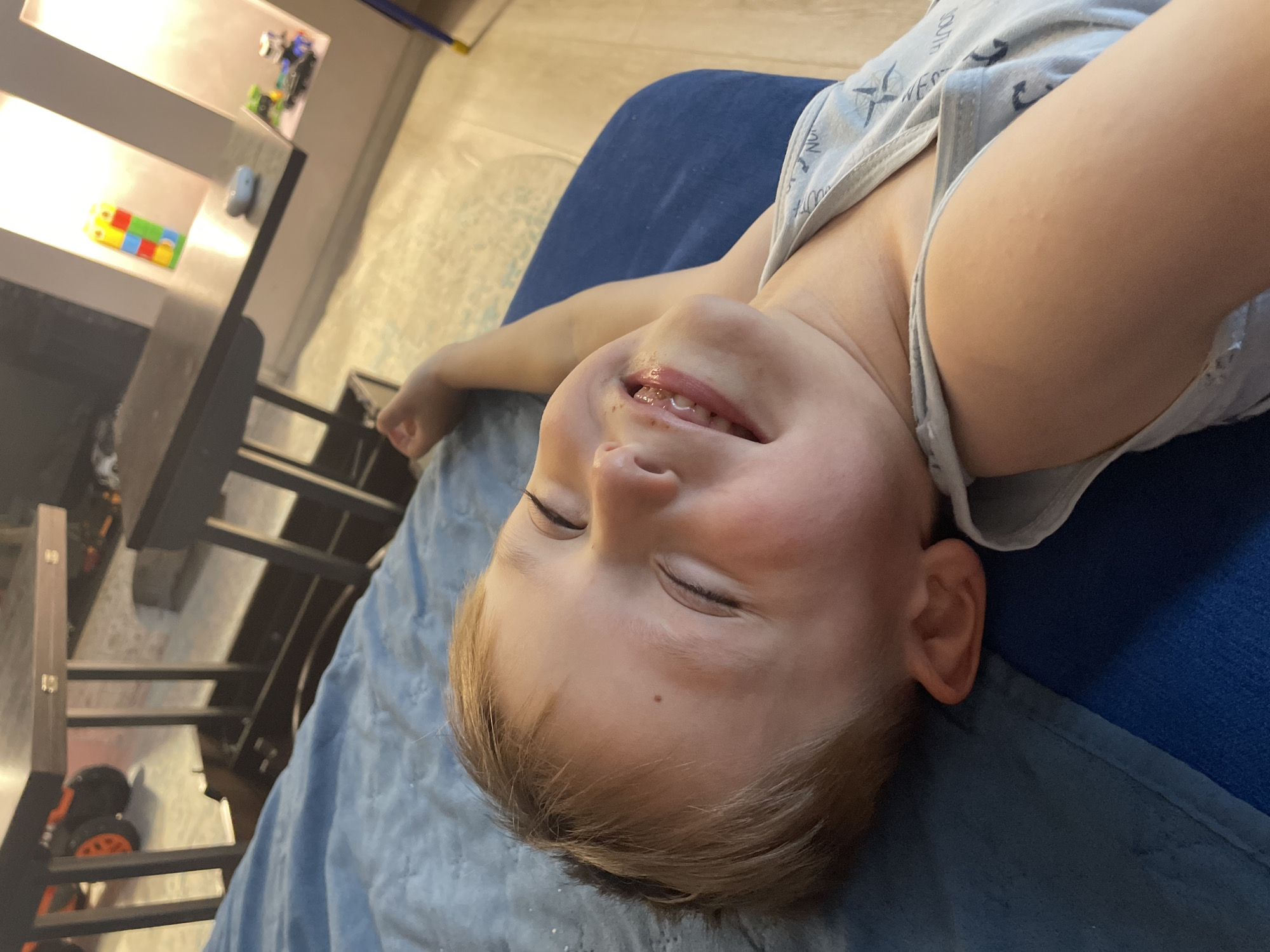 This media file depicts a close-up, head-on shot of a young child lying on a blue blanket. The child is on their back, with their head tilted back and eyes closed, a slight smile on their face, suggesting contentment or sleepiness. The child is wearing a light grey tank top with a faint pattern. Their hair is light brown and appears somewhat tousled.
In the background, out of focus, are elements of a domestic setting. To the left, there's a shelving unit with what appear to be toys, including a colorful block-like toy and some toy vehicles. Further back, a dark, possibly wooden, piece of furniture, like a desk or table, is visible. The flooring appears to be a light-colored surface. The lighting in the scene is soft, suggesting an indoor environment, possibly during daylight hours. The overall mood is peaceful and intimate, focusing on the child's relaxed state. There is no visible text within the frame.